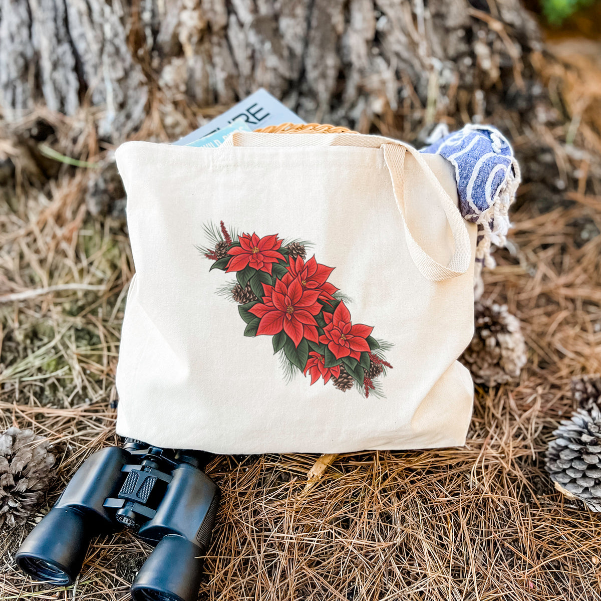 Canvas tote bag printed with a full-color illustration of red poinsettia flowers with pine greenery and pine cones, resting on dry grass beside a pair of binoculars.