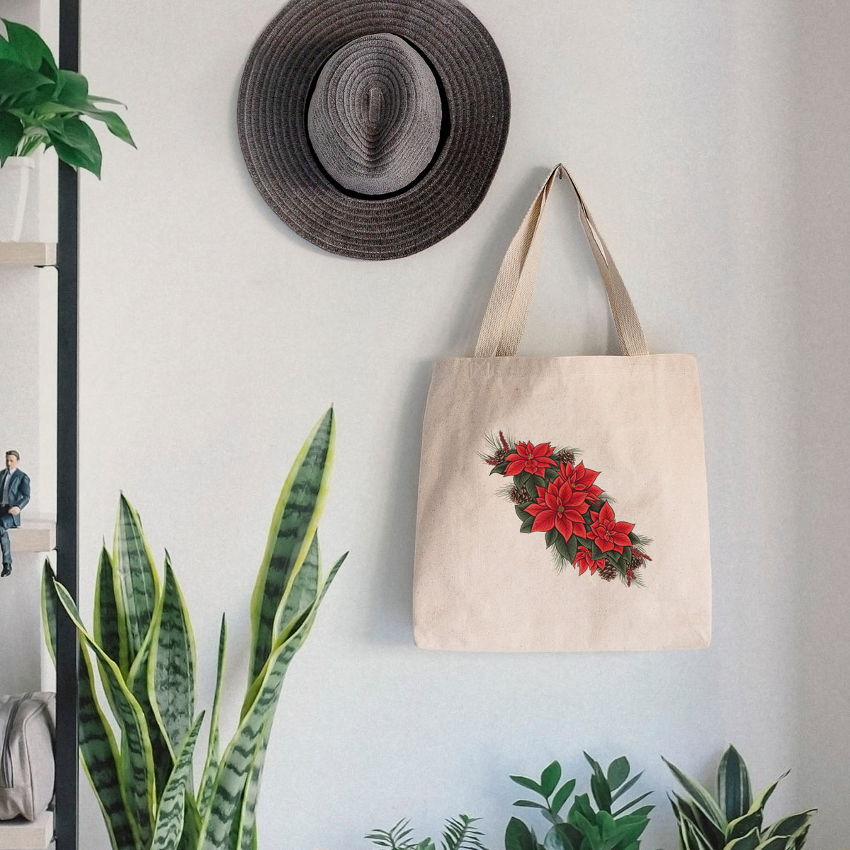 Canvas tote bag printed with a full-color illustration of red poinsettia flowers with pine greenery and pine cones, hanging on a wall hook below a hat, next to potted plants.