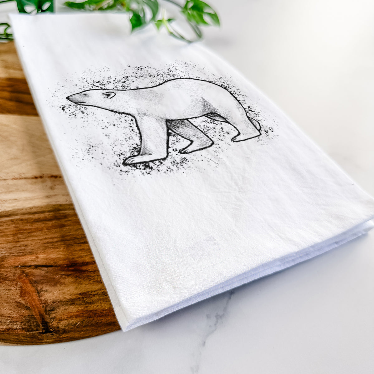 Close-up of a folded white tea towel on a wooden board, showing a black ink illustration of a polar bear walking across snow.