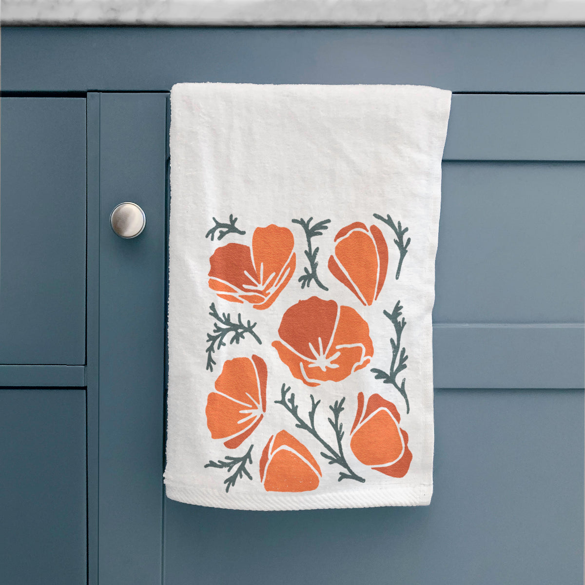 White hand towel printed with a full-color illustration of orange California poppies with blue-green leaves, hanging on a cabinet door.