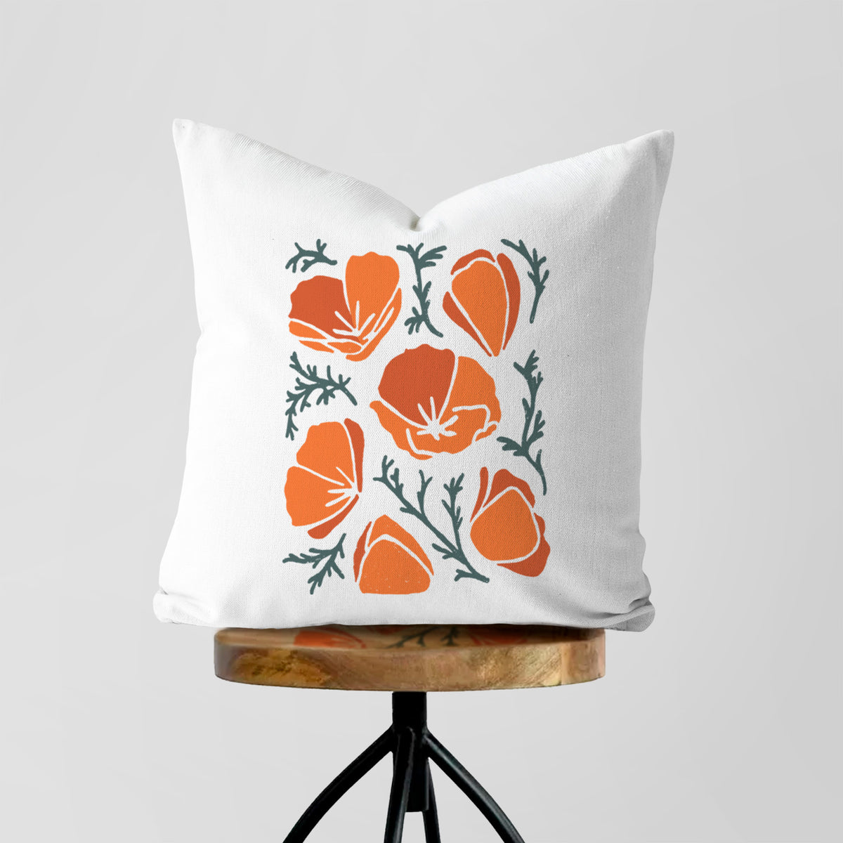 White pillow cover printed with a full-color illustration of orange California poppies with blue-green leaves, on a wooden stool.