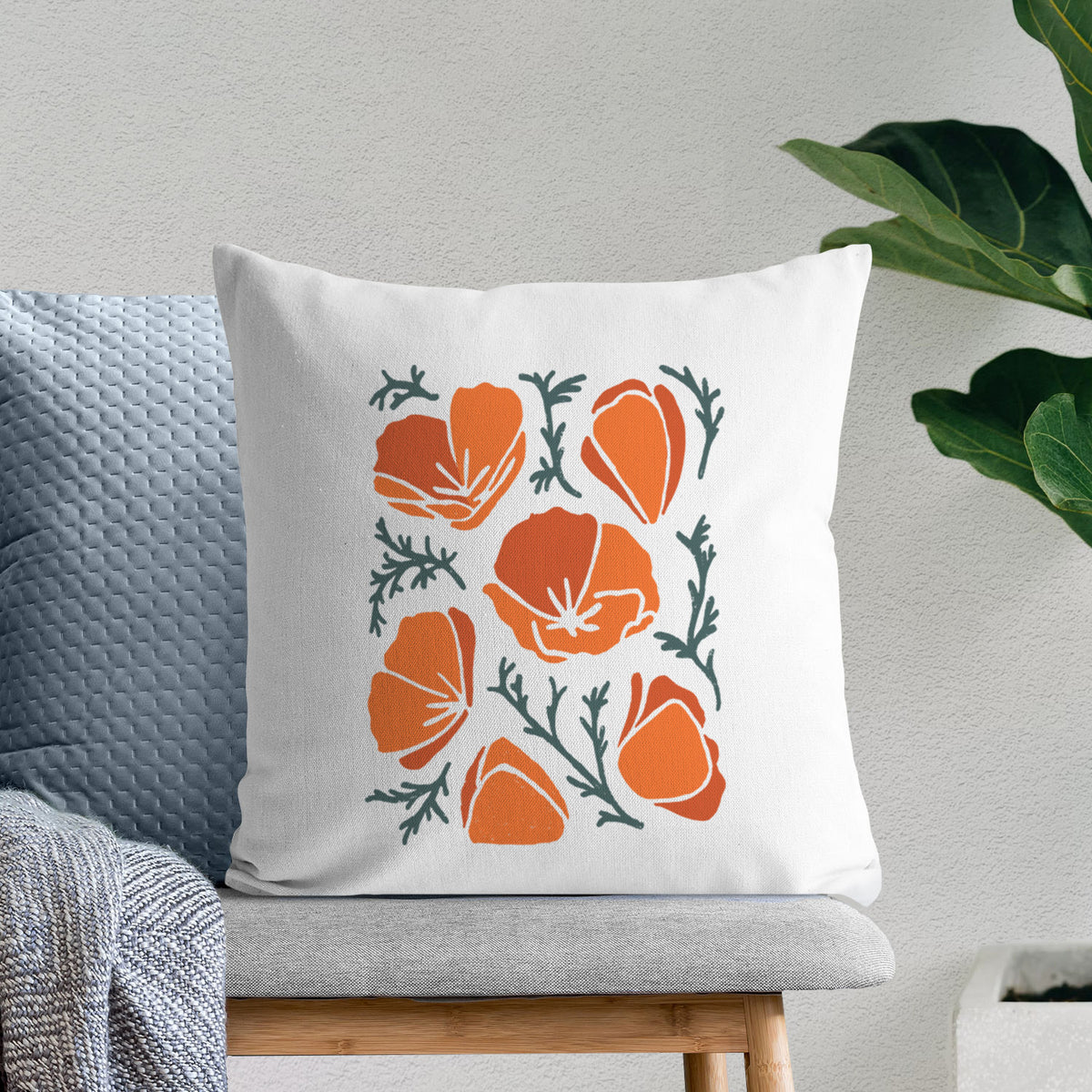 White pillow cover printed with a full-color illustration of orange California poppies with blue-green leaves, on a bench beside plants.