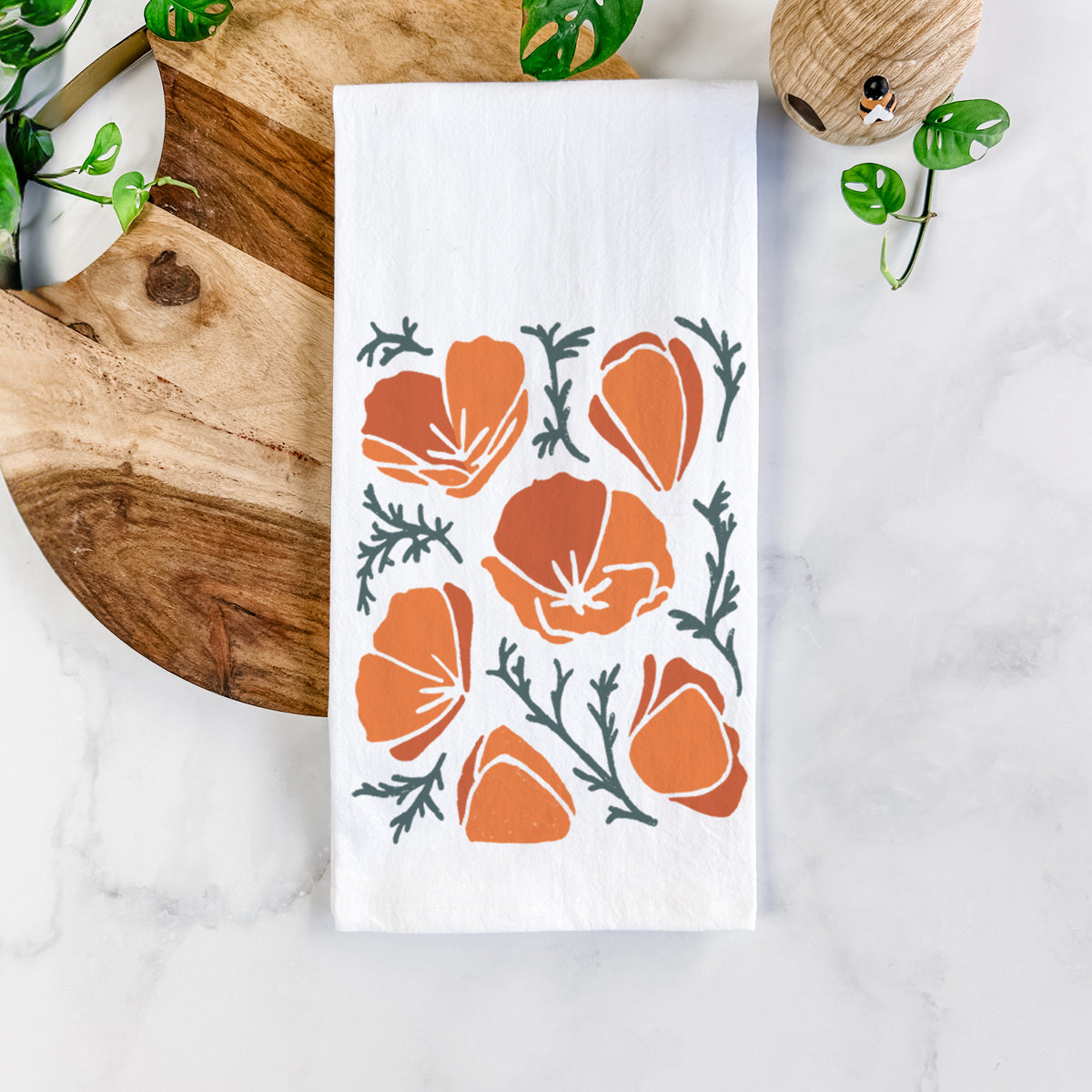 White cotton tea towel printed with a full-color illustration of orange California poppies with blue-green leaves, on a marble counter beside a round wooden cutting board and greenery.