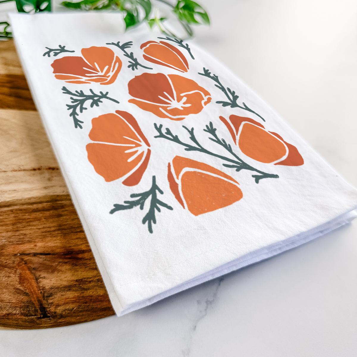 Close-up of a folded white tea towel on a wooden board, showing a full-color illustration of orange California poppies with blue-green leaves.