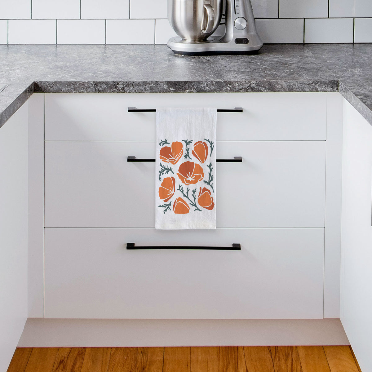 White tea towel printed with a full-color illustration of orange California poppies with blue-green leaves, hanging from a drawer handle in a bright white kitchen.