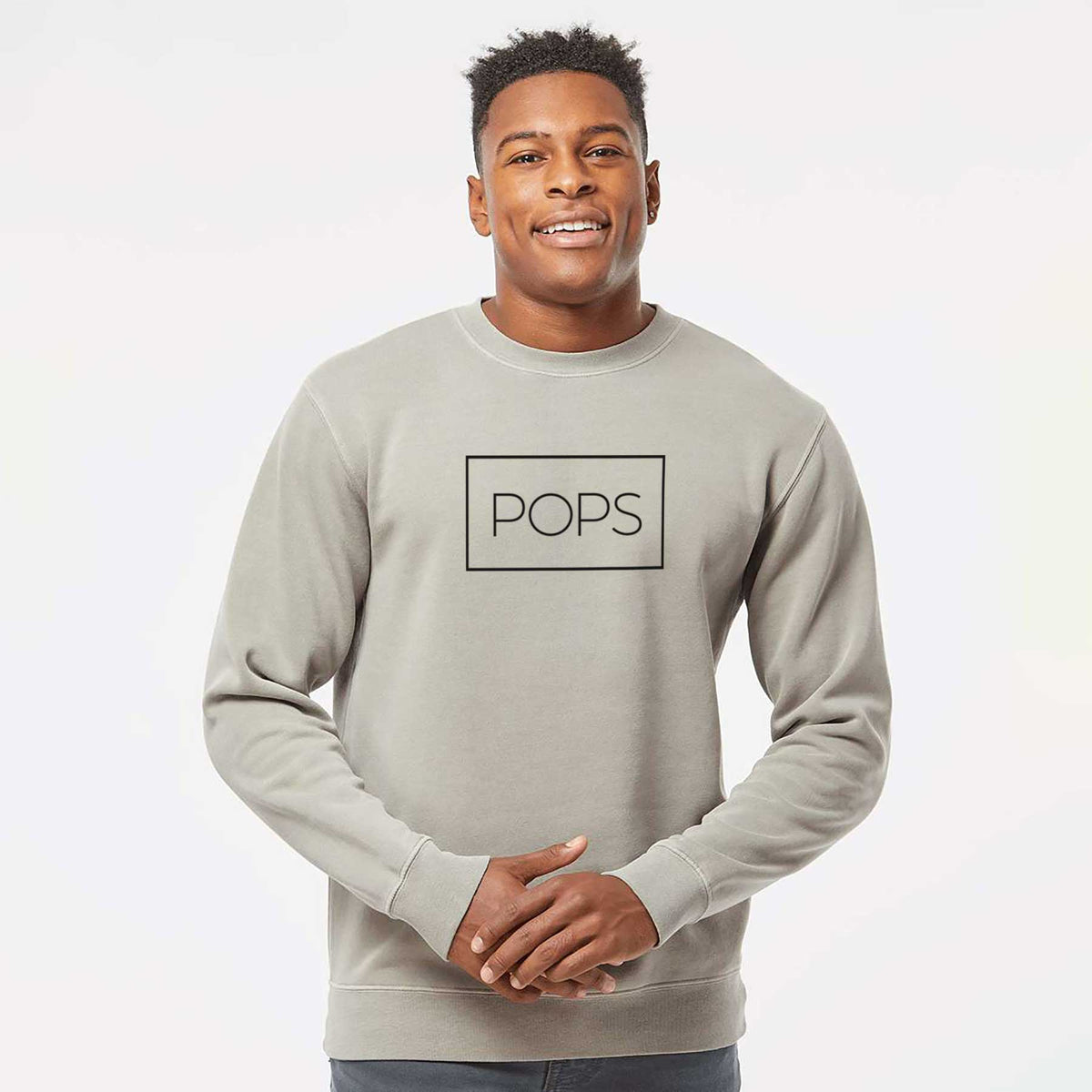 Model wearing a cement grey pigment dyed crewneck sweatshirt printed with the word POPS in thin black letters inside a rectangle outline.