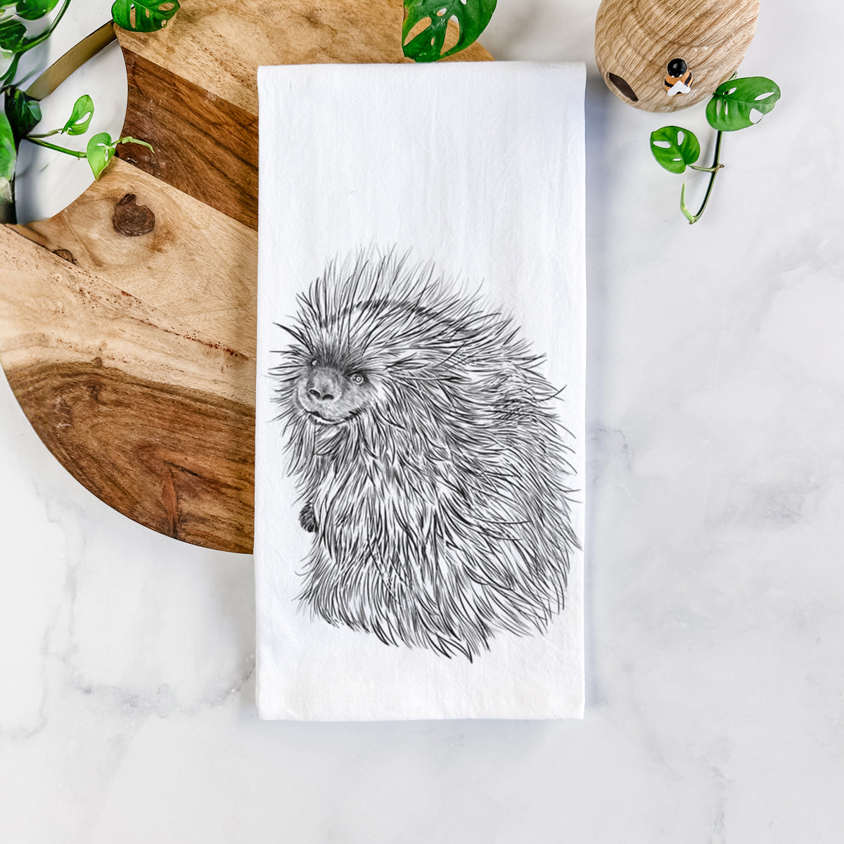 White cotton tea towel printed with a detailed black ink illustration of a porcupine with its quills raised, on a marble counter beside a round wooden cutting board and greenery.