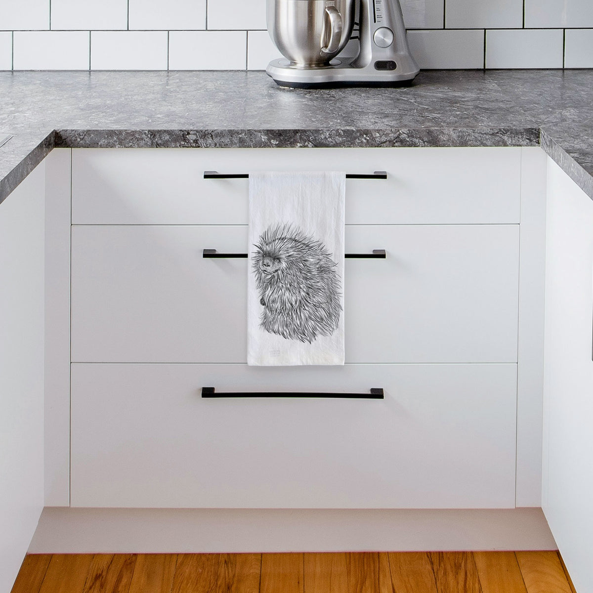 White tea towel printed with a detailed black ink illustration of a porcupine with its quills raised, hanging from a drawer handle in a bright white kitchen.