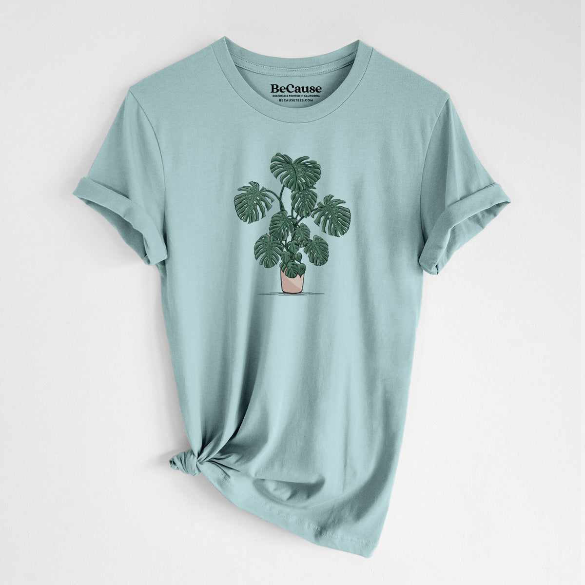 Dusty blue lightweight cotton t-shirt laid flat with the sleeves rolled, printed with a full-color illustration of a large potted Monstera plant.
