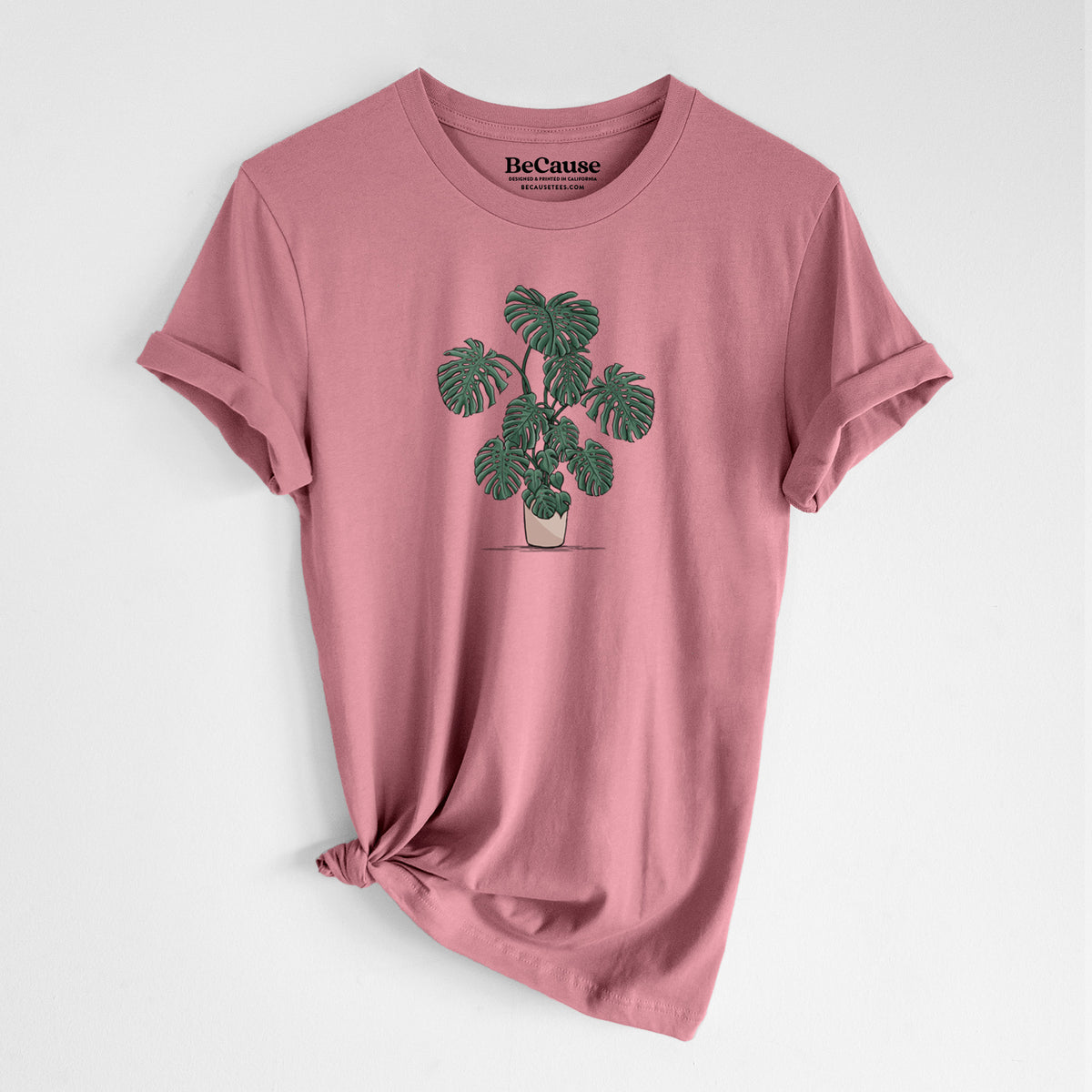 Mauve pink lightweight cotton t-shirt laid flat with the sleeves rolled, printed with a full-color illustration of a large potted Monstera plant.