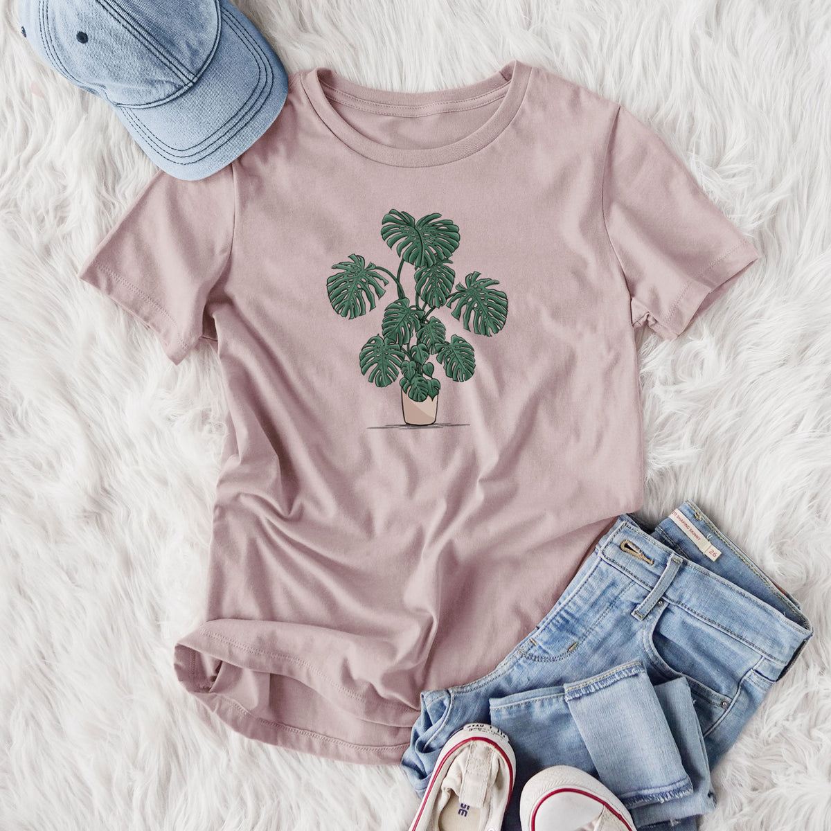 Women's relaxed fit cotton t-shirt printed with a full-color illustration of a large potted Monstera plant, styled flat on a white faux fur rug with jeans.