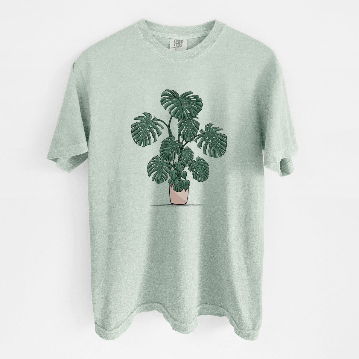Bay green Comfort Colors t-shirt laid flat, printed with a full-color illustration of a large potted Monstera plant.