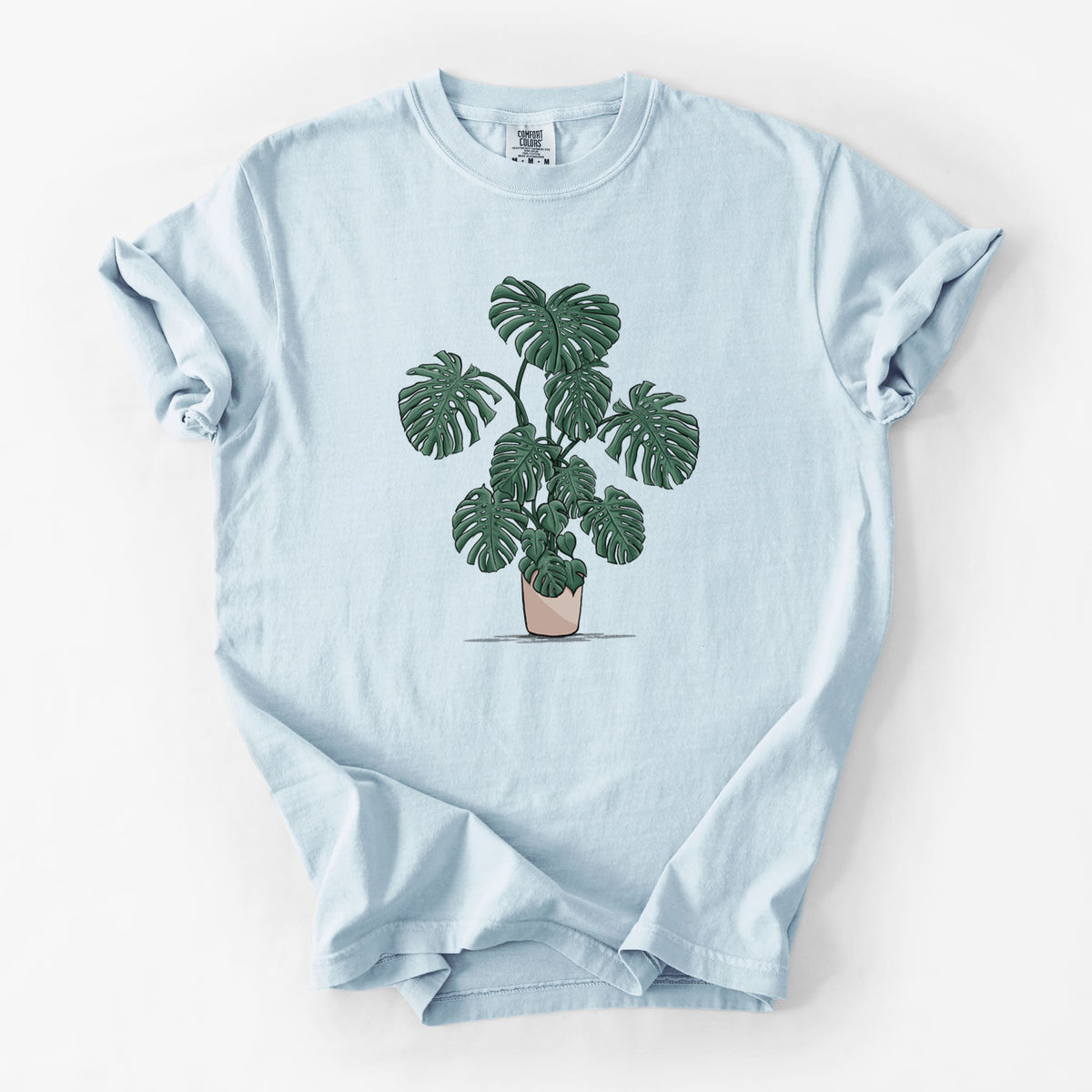 Chambray blue Comfort Colors t-shirt laid flat, printed with a full-color illustration of a large potted Monstera plant.