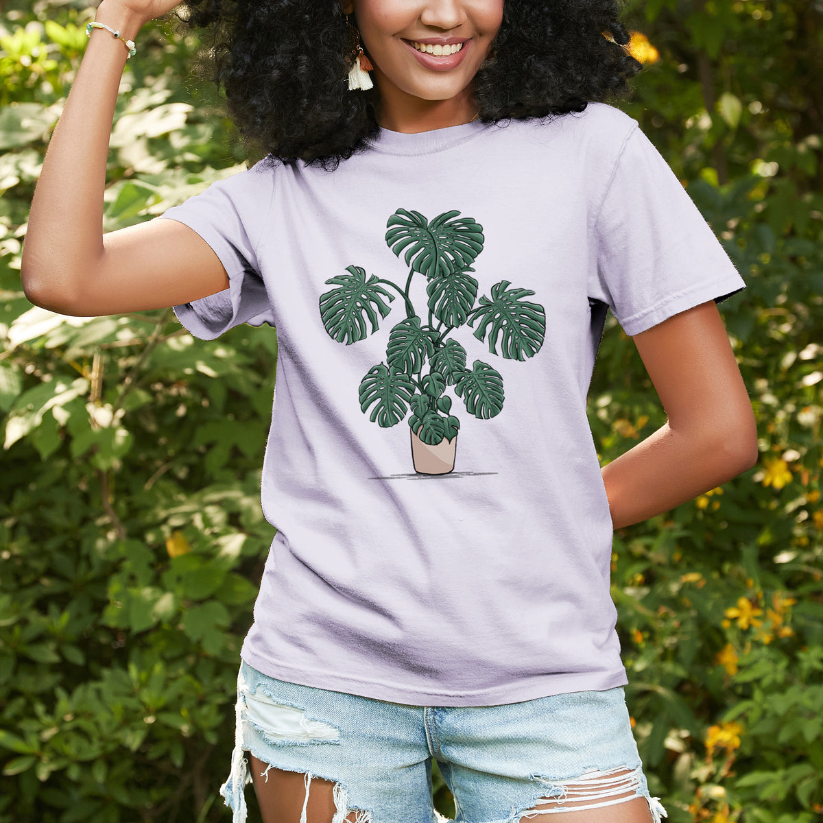 Woman wearing a Comfort Colors t-shirt printed with a full-color illustration of a large potted Monstera plant, outdoors.