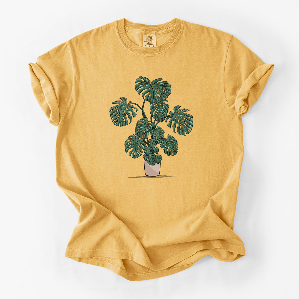 Golden wheat yellow Comfort Colors t-shirt laid flat, printed with a full-color illustration of a large potted Monstera plant.