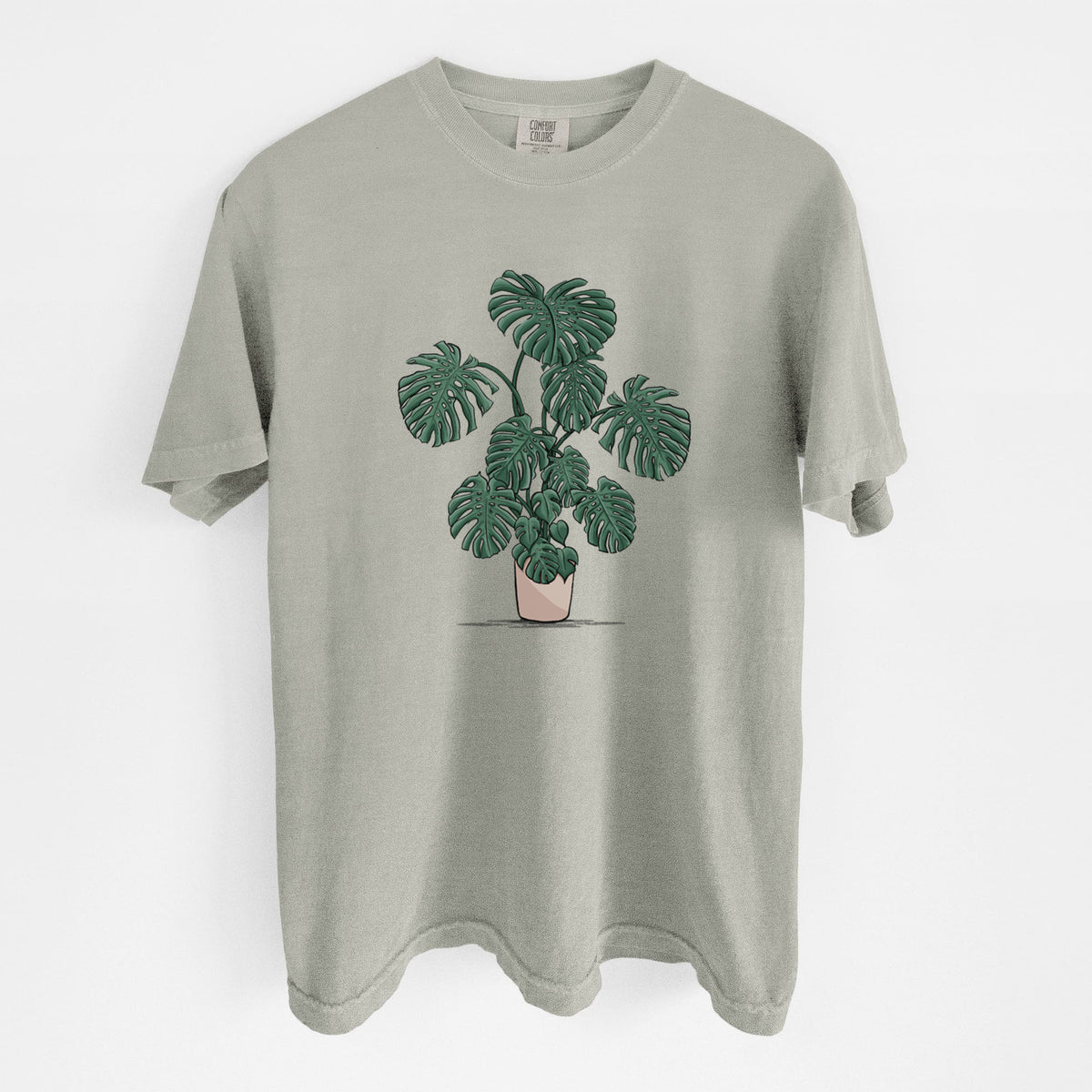 Sandstone beige Comfort Colors t-shirt laid flat, printed with a full-color illustration of a large potted Monstera plant.