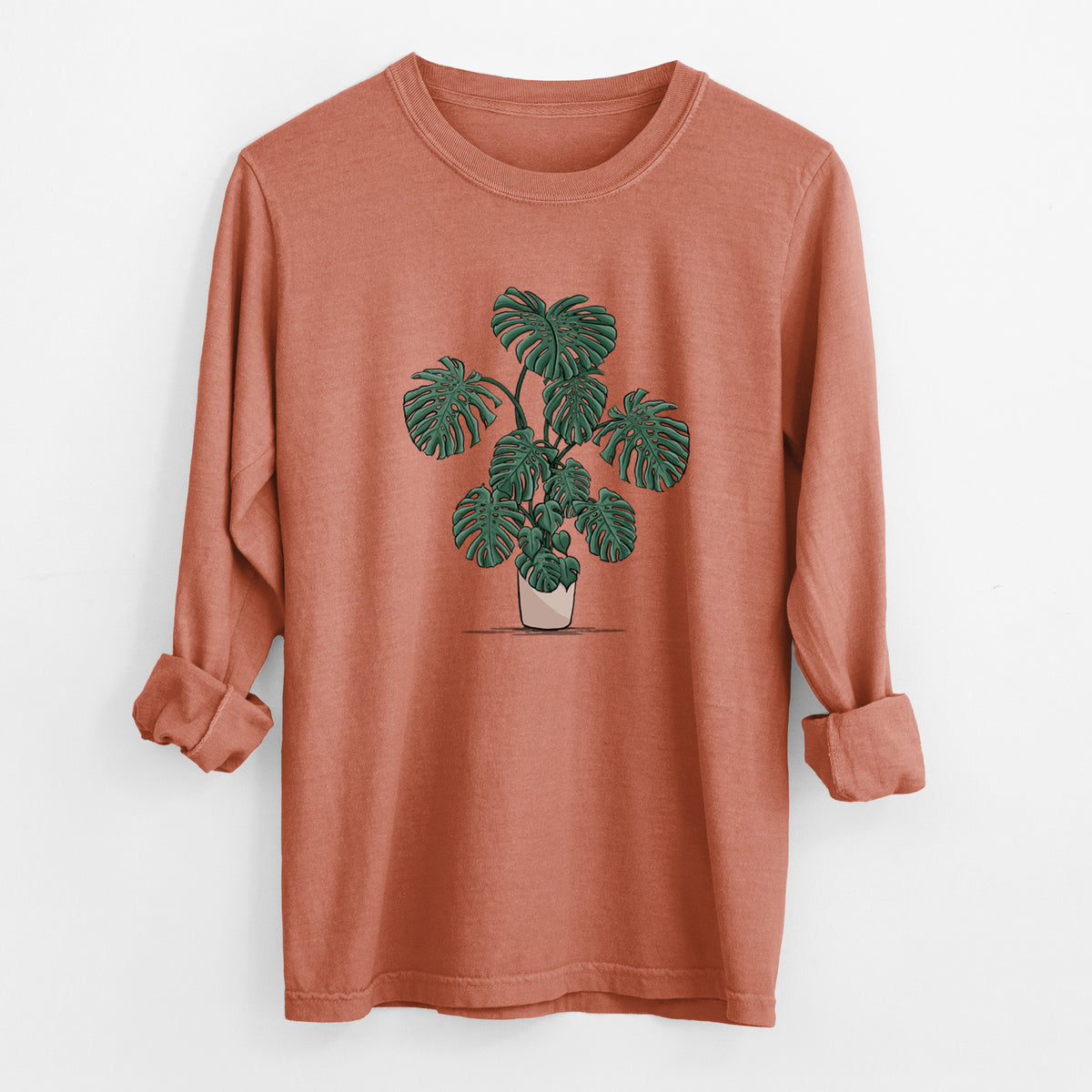 Yam burnt orange Comfort Colors long sleeve shirt laid flat with the cuffs rolled, printed with a full-color illustration of a large potted Monstera plant.