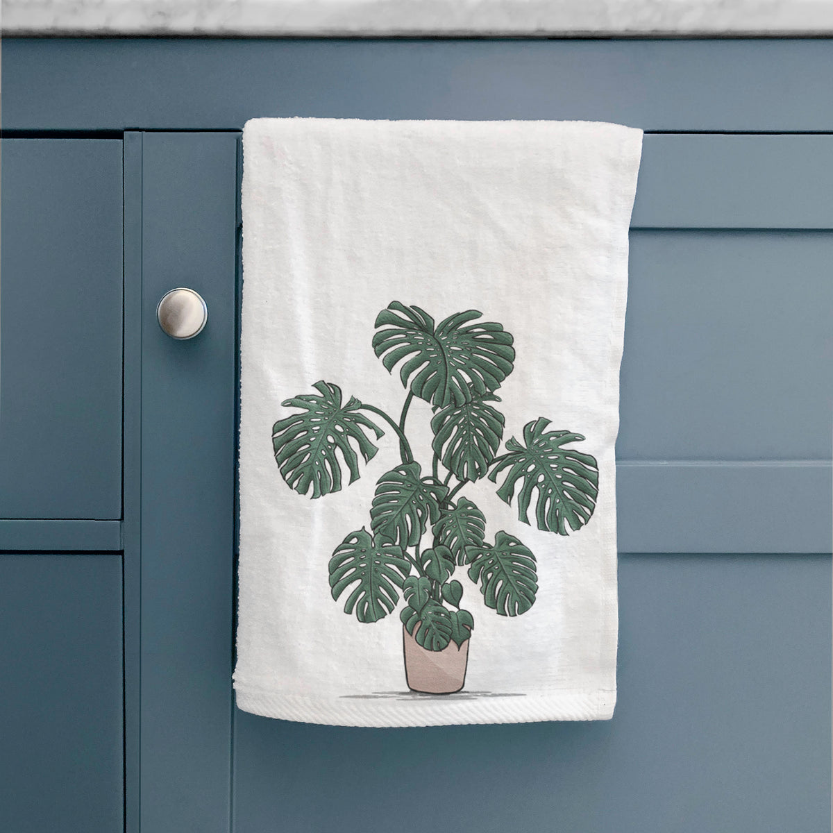 White hand towel printed with a full-color illustration of a large potted Monstera plant, hanging on a cabinet door.
