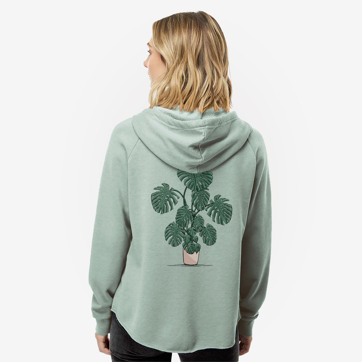 Woman wearing a sage green women's Cali Wave zip-up sweatshirt printed with a full-color illustration of a large potted Monstera plant, seen from behind.