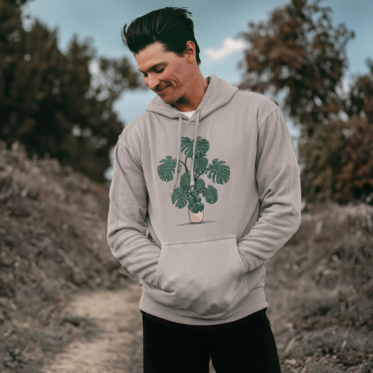 Man wearing a cement grey pigment dyed hoodie printed with a full-color illustration of a large potted Monstera plant.
