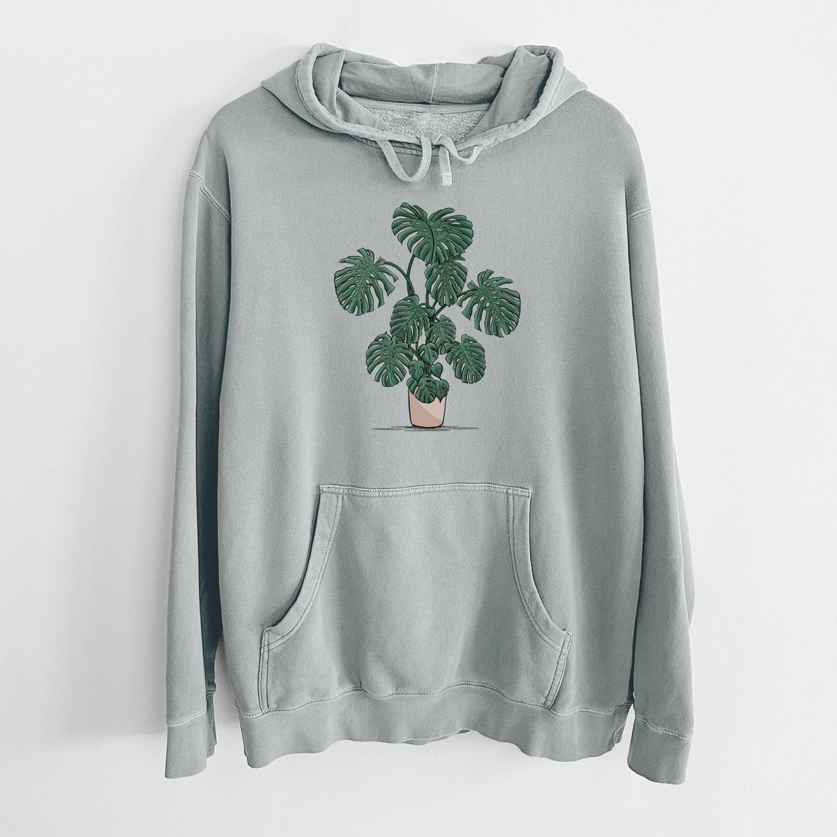 Sage green pigment dyed hoodie laid flat, printed with a full-color illustration of a large potted Monstera plant.