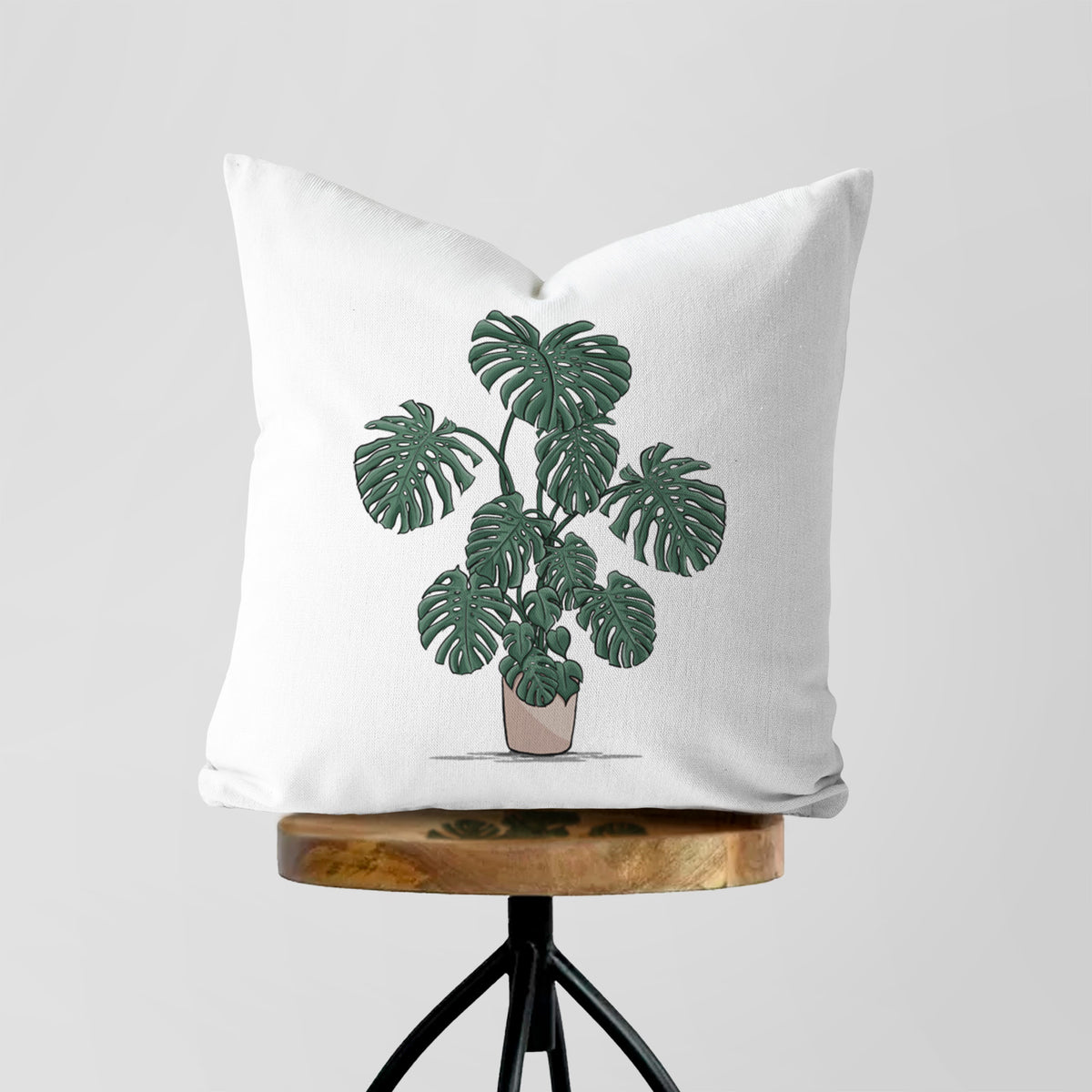 White pillow cover printed with a full-color illustration of a large potted Monstera plant, on a wooden stool.