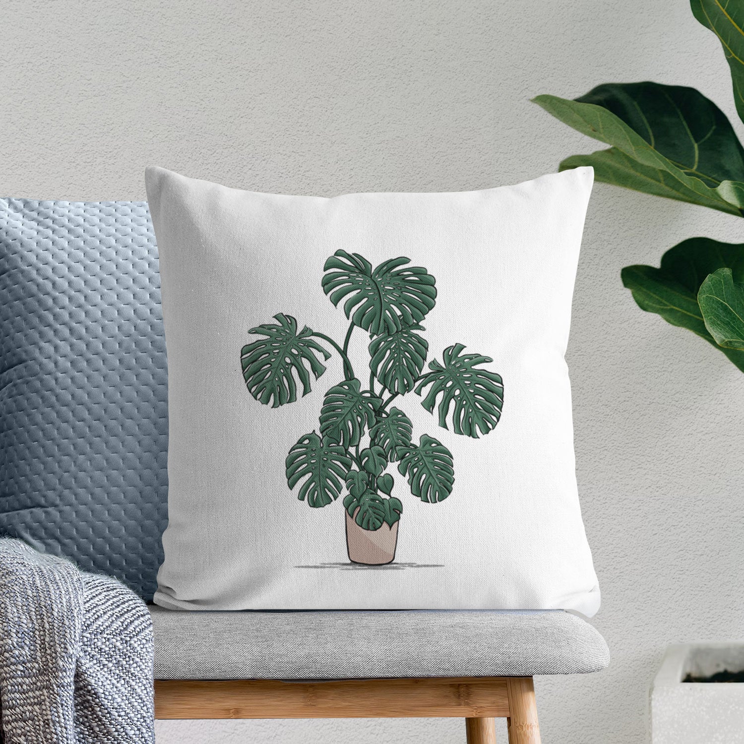 White pillow cover printed with a full-color illustration of a large potted Monstera plant, on a bench beside plants.