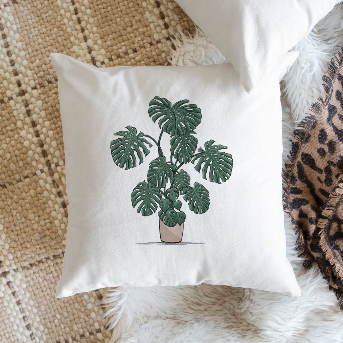 White pillow cover printed with a full-color illustration of a large potted Monstera plant, on a woven chair.