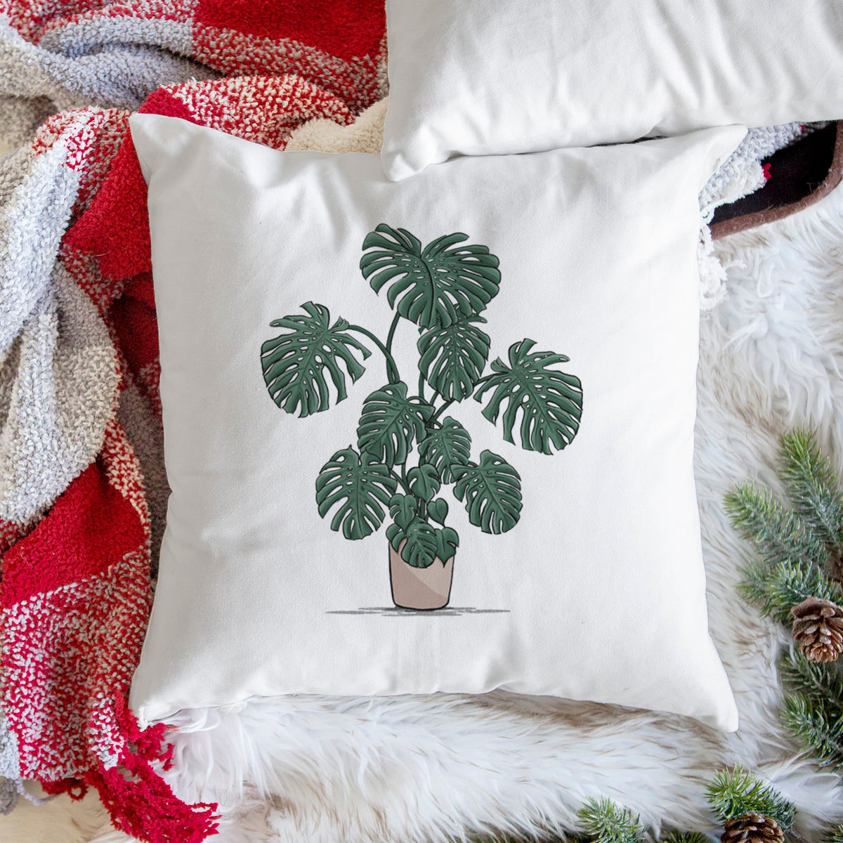 White pillow cover printed with a full-color illustration of a large potted Monstera plant, on a woven chair.