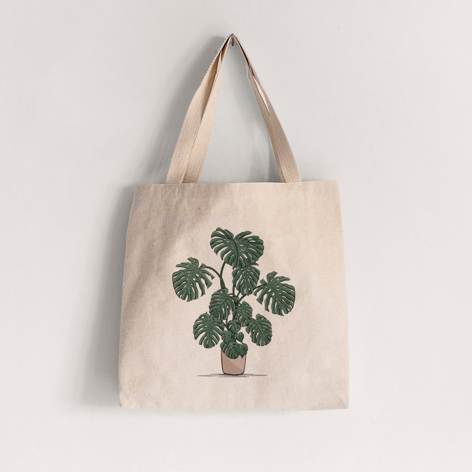 Natural canvas tote bag printed with a full-color illustration of a large potted Monstera plant.