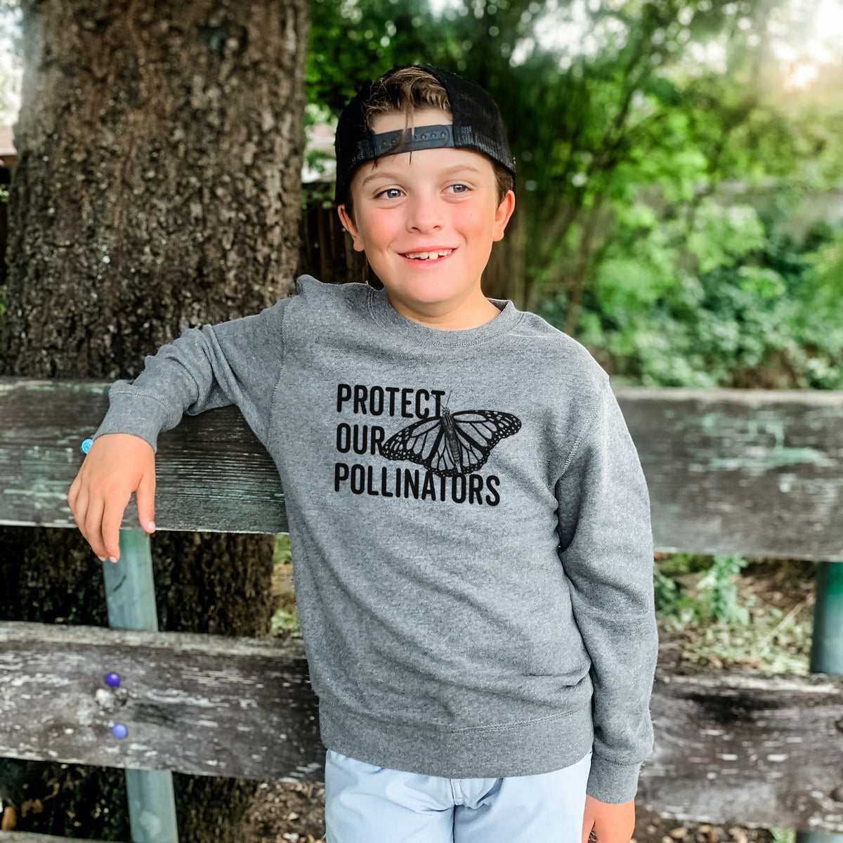 Boy wearing a youth crewneck sweatshirt printed with bold black words reading Protect Our Pollinators beside a monarch butterfly, outdoors.