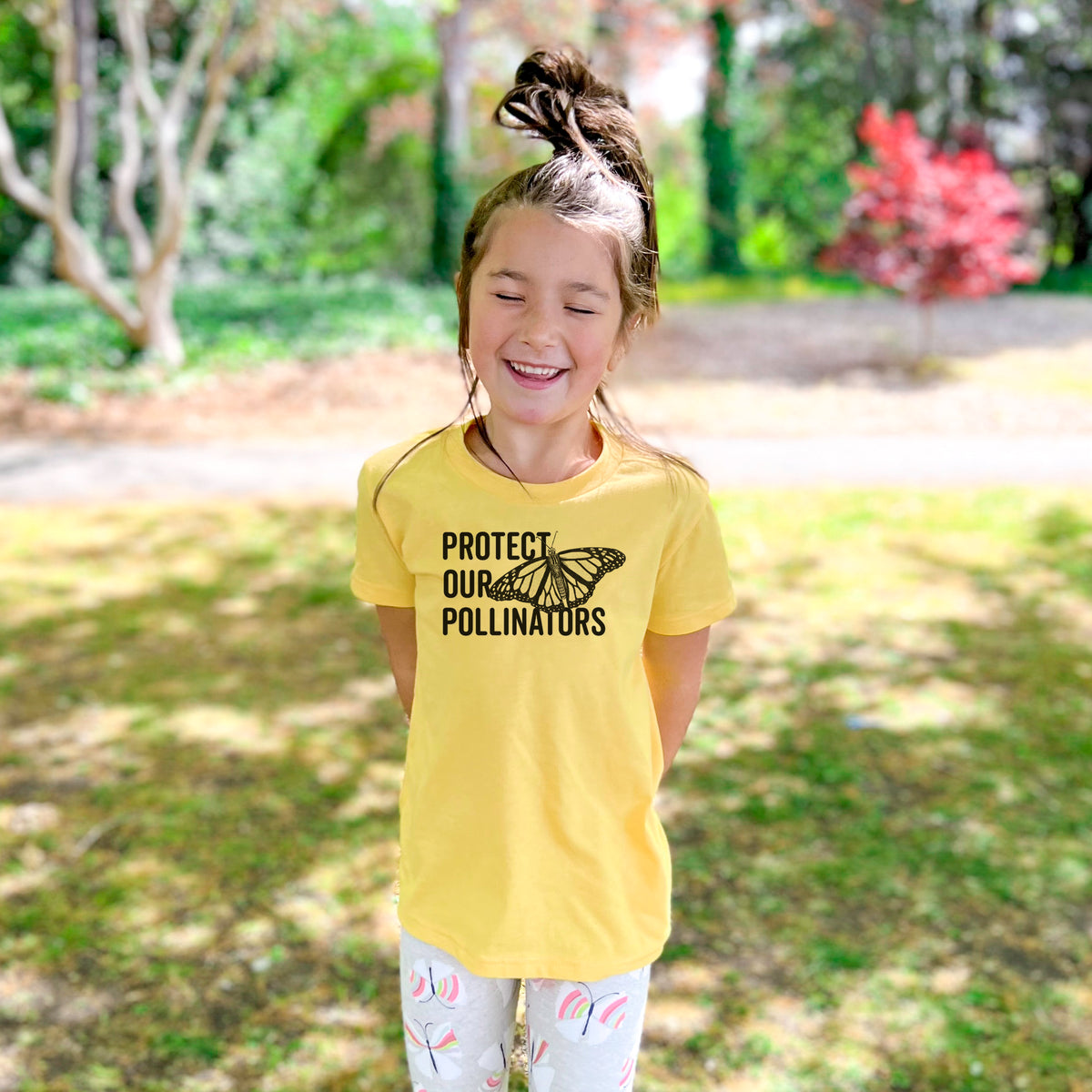 Smiling girl wearing a youth t-shirt printed with bold black words reading Protect Our Pollinators beside a monarch butterfly, outdoors.