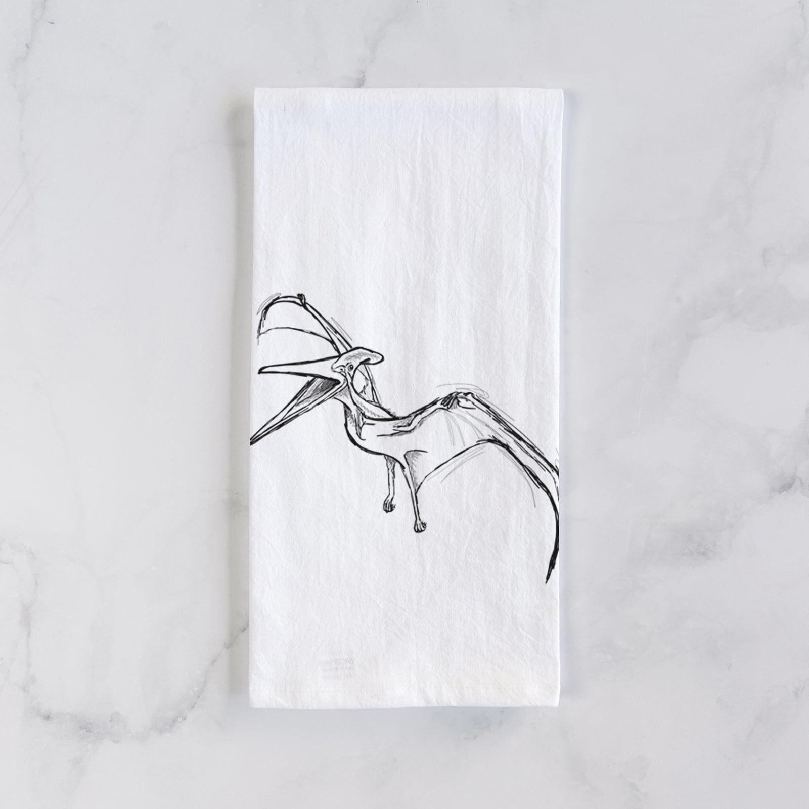 White cotton tea towel printed with a black ink illustration of a Pteranodon in flight, on a marble counter beside a round wooden cutting board and greenery.
