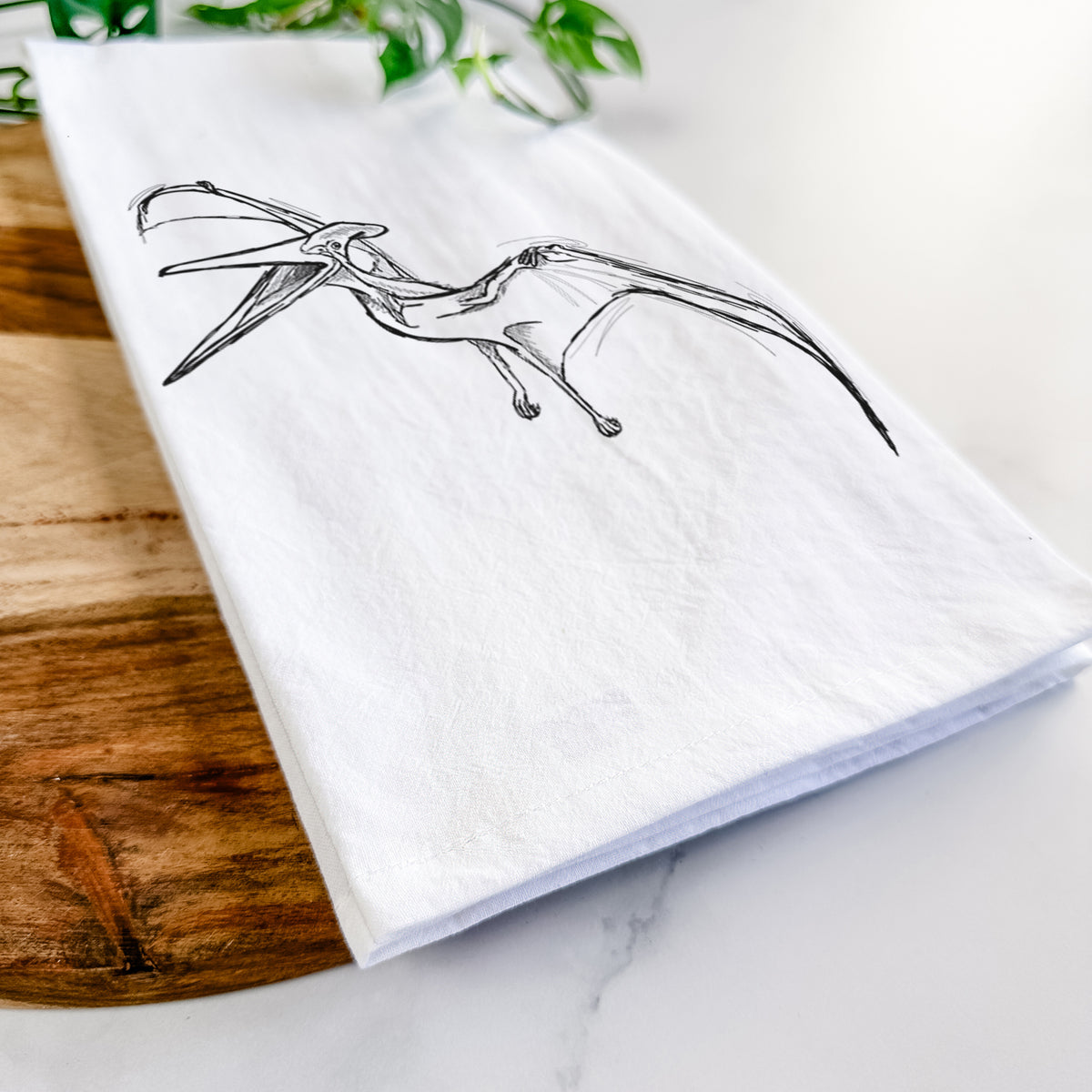 Close-up of a folded white tea towel on a wooden board, showing a black ink illustration of a Pteranodon in flight.