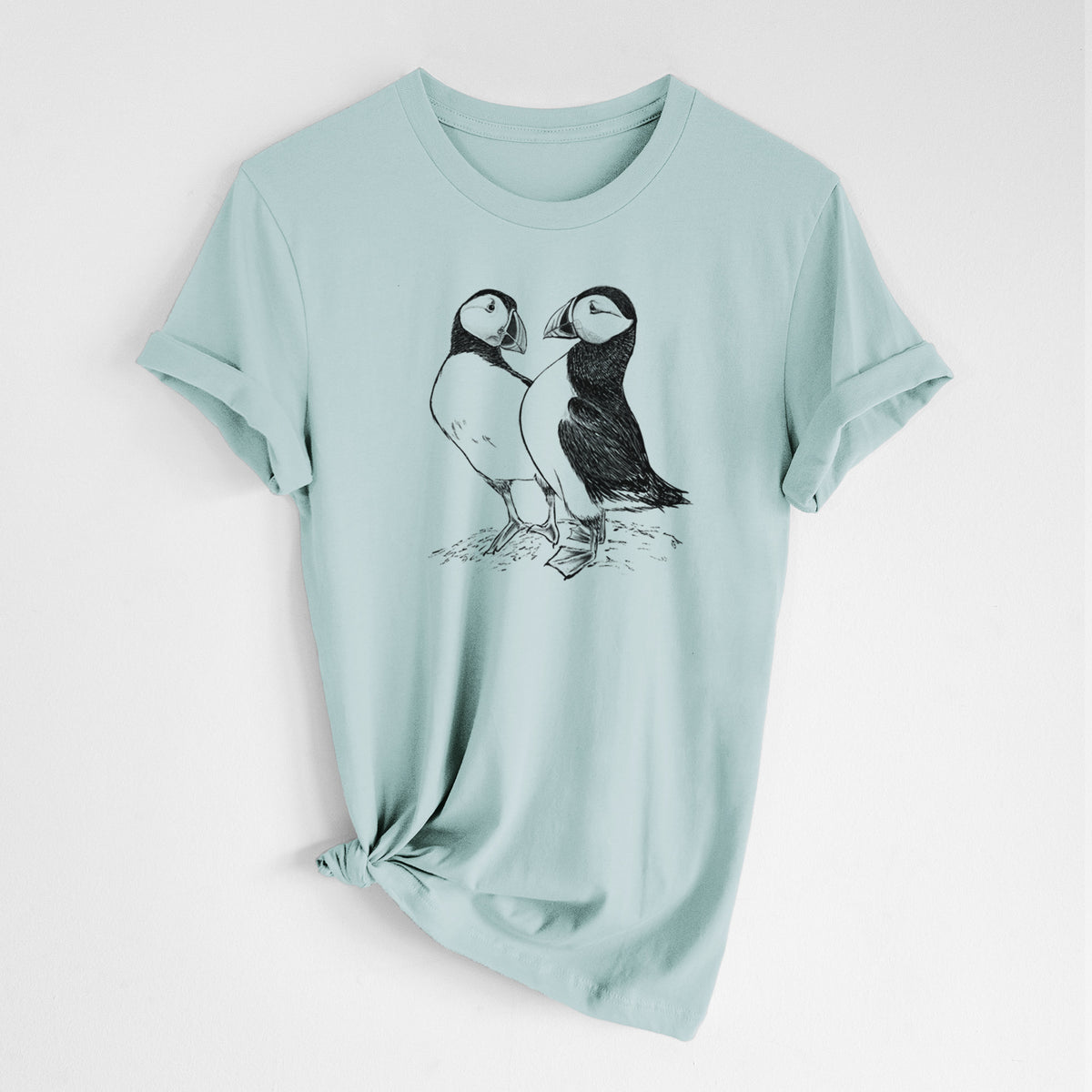 Dusty blue lightweight cotton t-shirt laid flat with the sleeves rolled, printed with a detailed black ink illustration of two Atlantic puffins standing side by side.