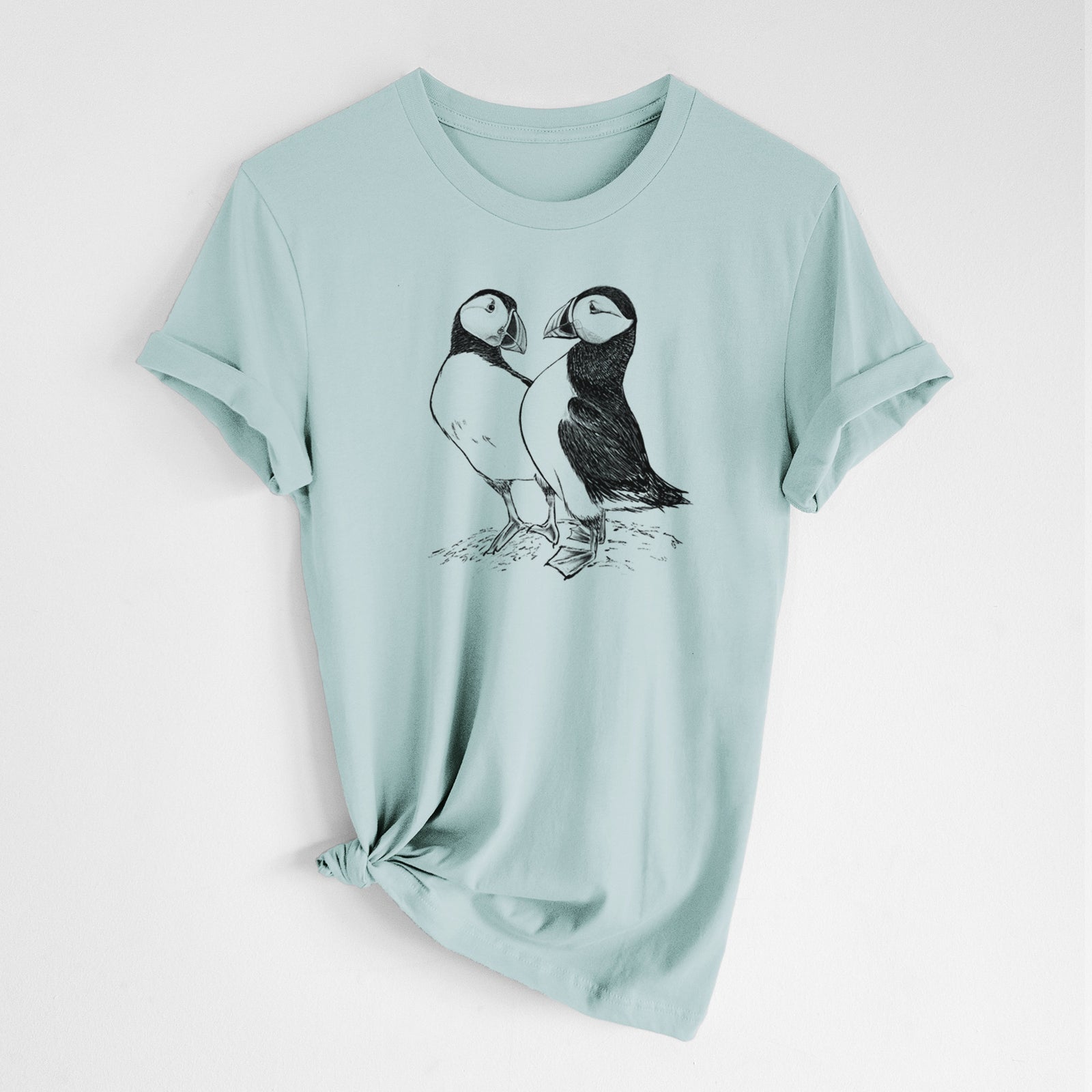 Dusty blue lightweight cotton t-shirt laid flat with the sleeves rolled, printed with a detailed black ink illustration of two Atlantic puffins standing side by side.
