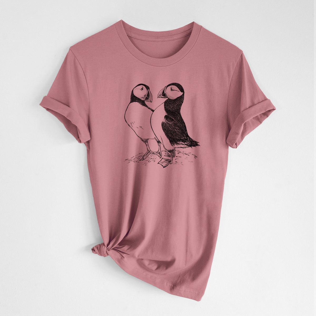 Mauve pink lightweight cotton t-shirt laid flat with the sleeves rolled, printed with a detailed black ink illustration of two Atlantic puffins standing side by side.