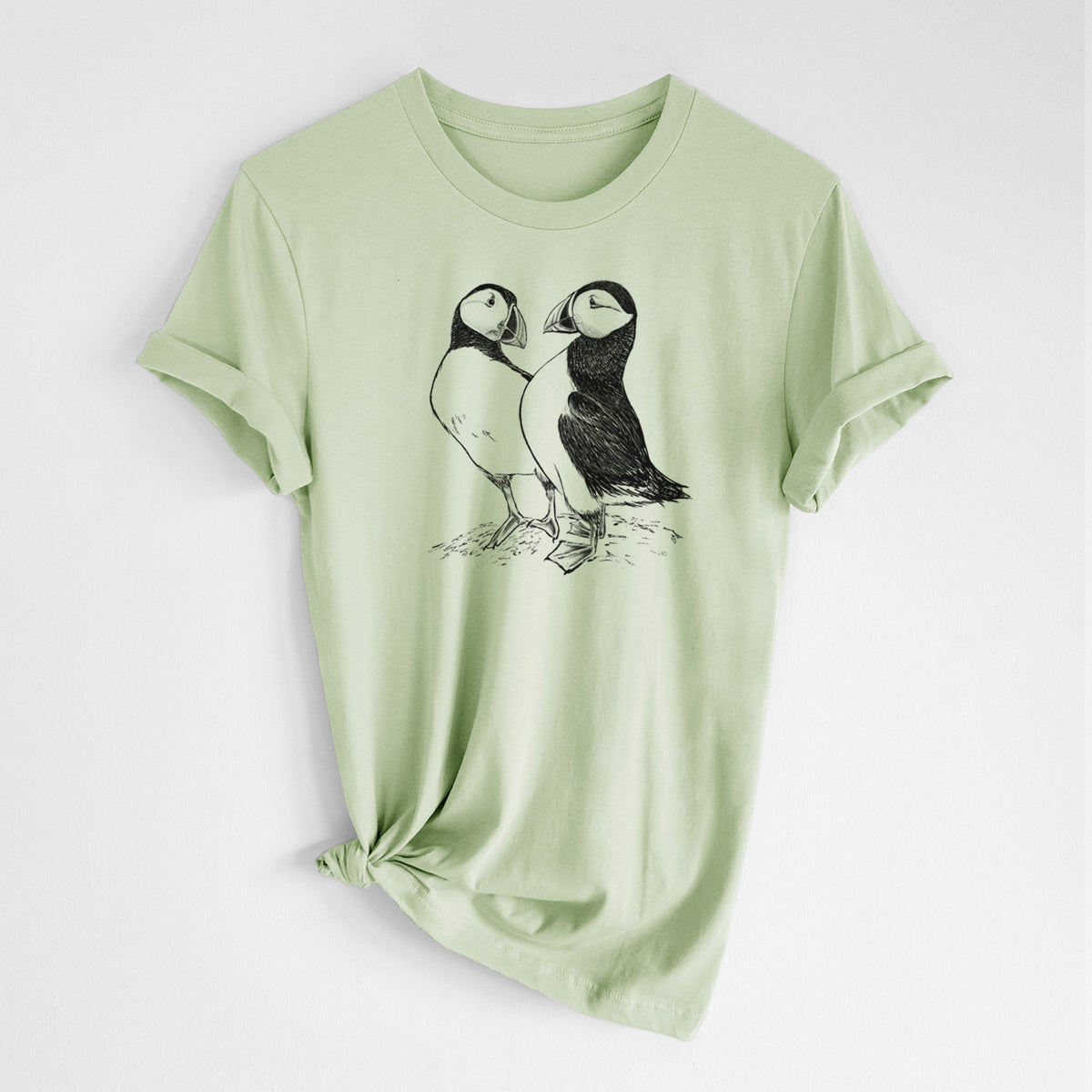 Spring green lightweight cotton t-shirt laid flat with the sleeves rolled, printed with a detailed black ink illustration of two Atlantic puffins standing side by side.