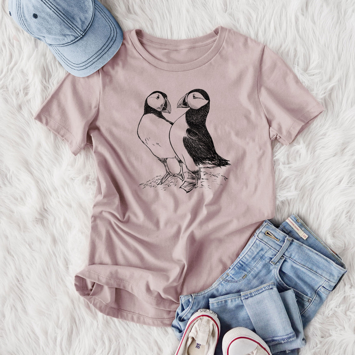 Women's relaxed fit cotton t-shirt printed with a detailed black ink illustration of two Atlantic puffins standing side by side, styled flat on a white faux fur rug with jeans.