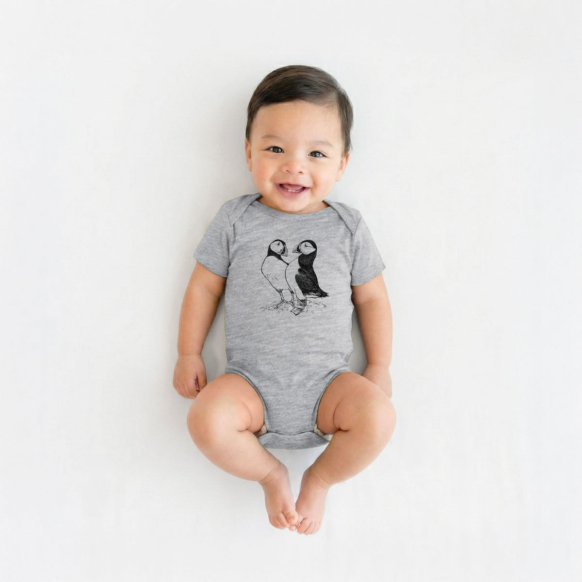 Smiling baby boy wearing a grey baby bodysuit printed with a detailed black ink illustration of two Atlantic puffins standing side by side.