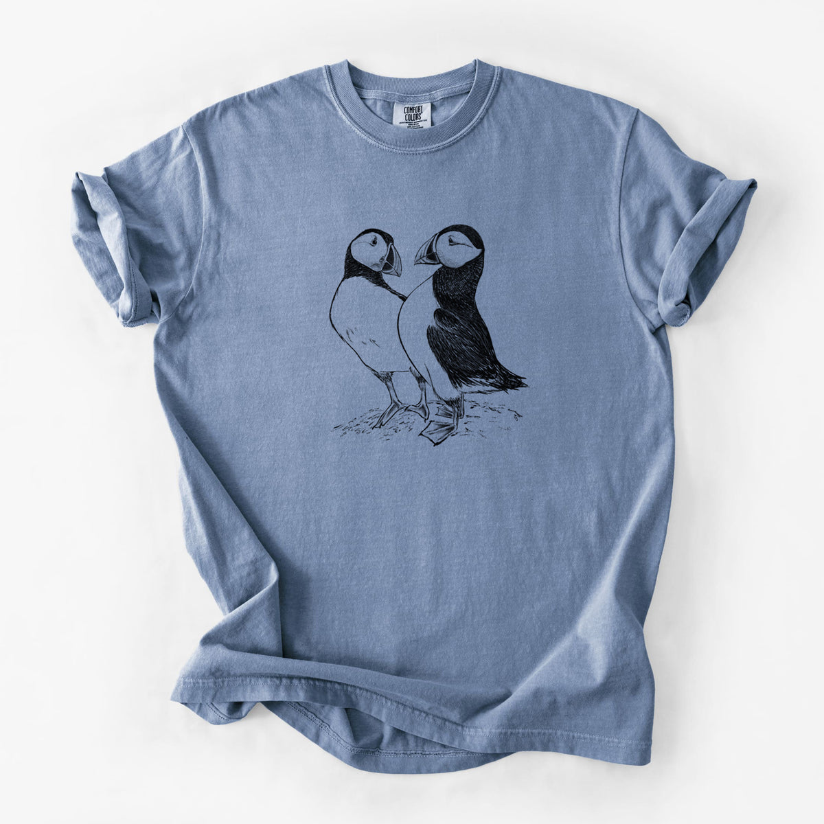 Blue jean Comfort Colors t-shirt laid flat, printed with a detailed black ink illustration of two Atlantic puffins standing side by side.