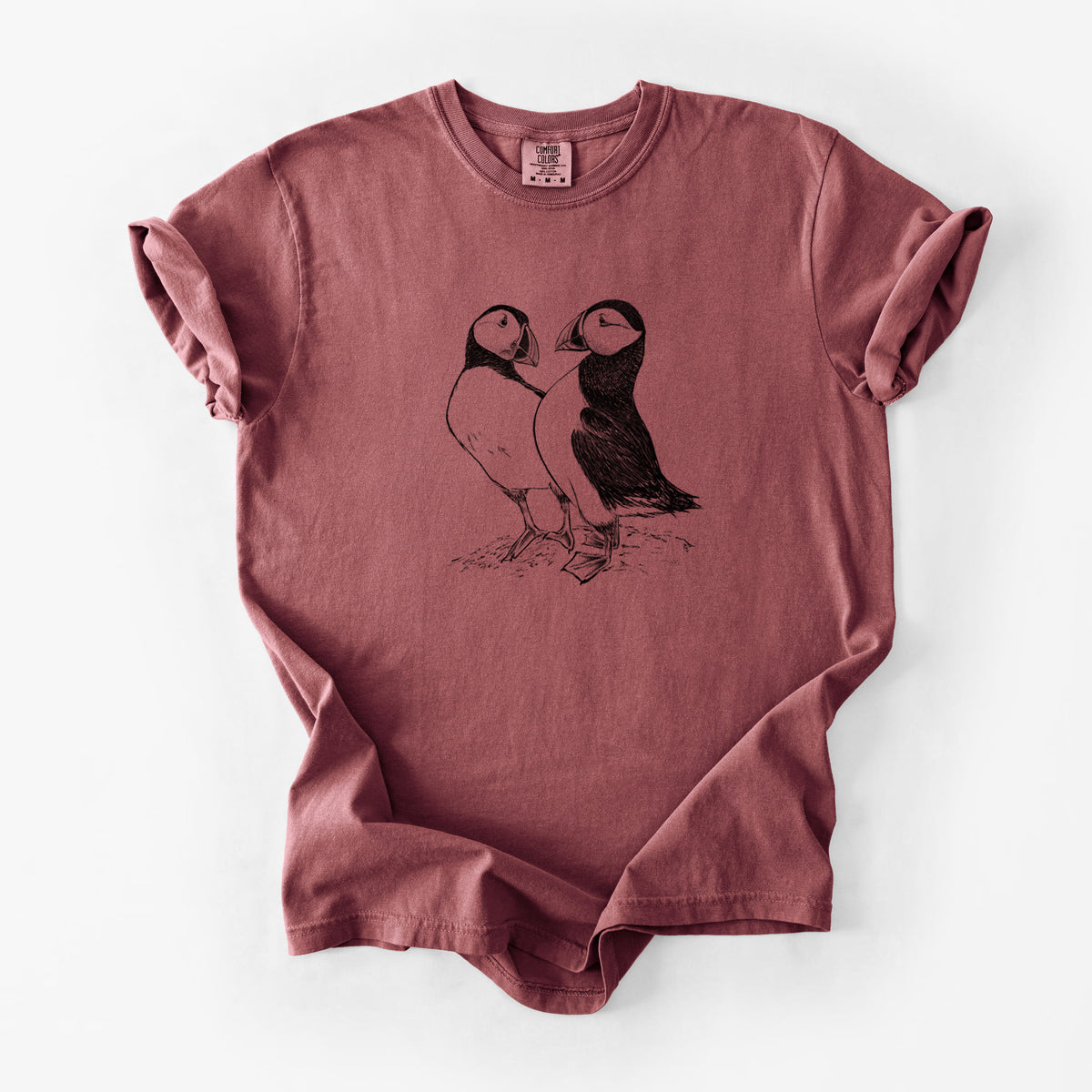 Brick red Comfort Colors t-shirt laid flat, printed with a detailed black ink illustration of two Atlantic puffins standing side by side.