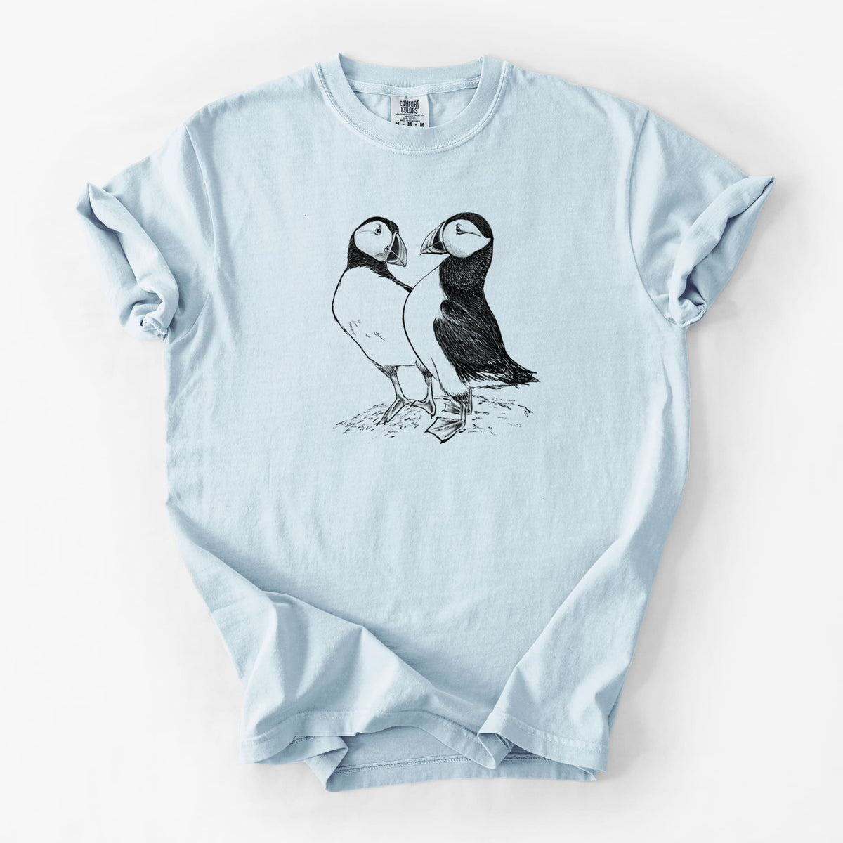 Chambray blue Comfort Colors t-shirt laid flat, printed with a detailed black ink illustration of two Atlantic puffins standing side by side.
