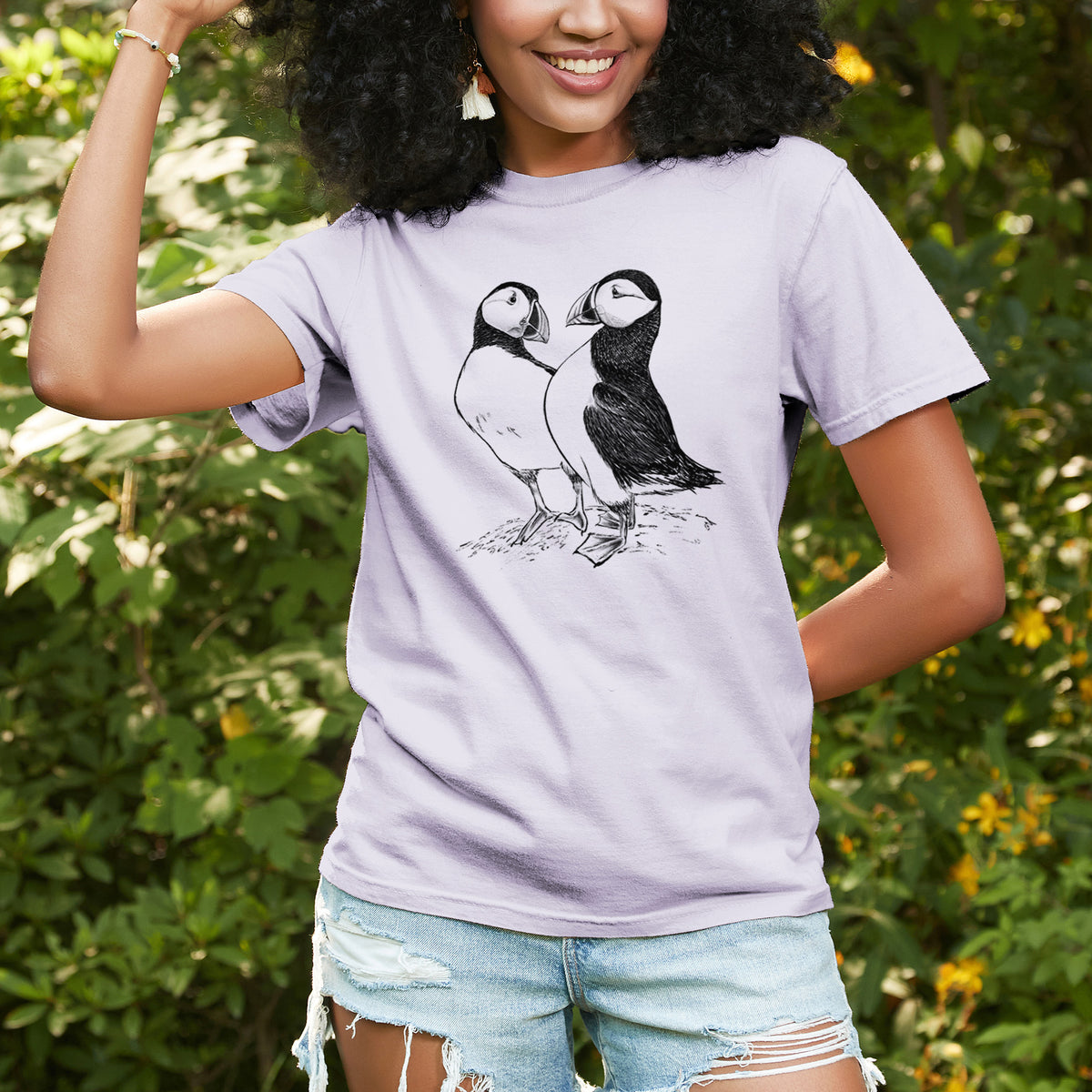 Woman wearing a Comfort Colors t-shirt printed with a detailed black ink illustration of two Atlantic puffins standing side by side, outdoors.
