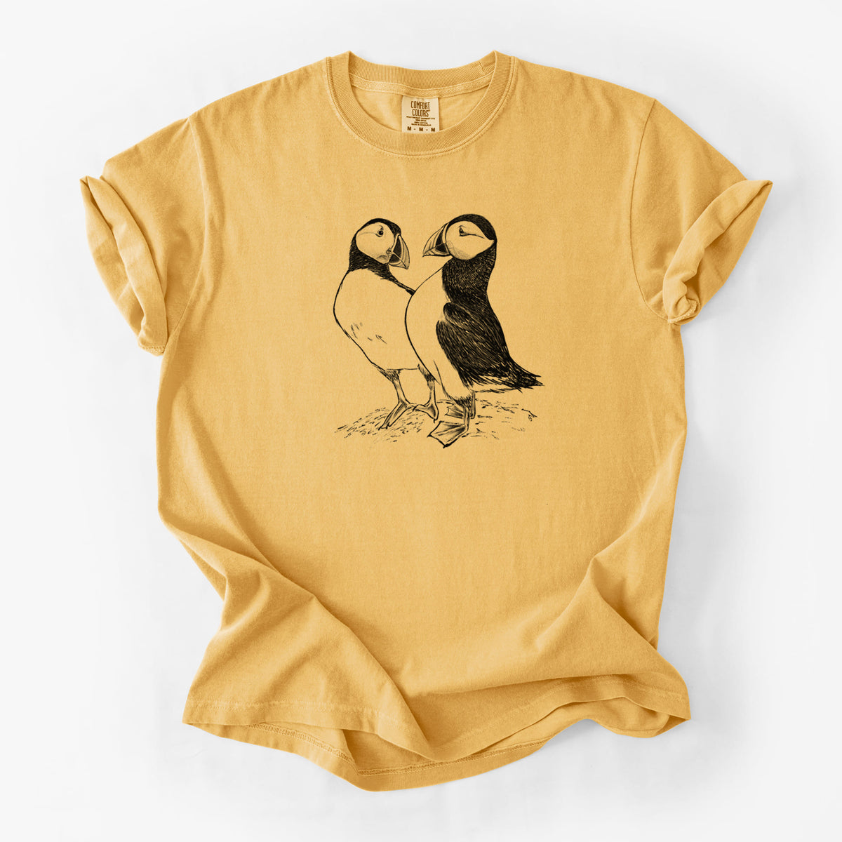Golden wheat yellow Comfort Colors t-shirt laid flat, printed with a detailed black ink illustration of two Atlantic puffins standing side by side.