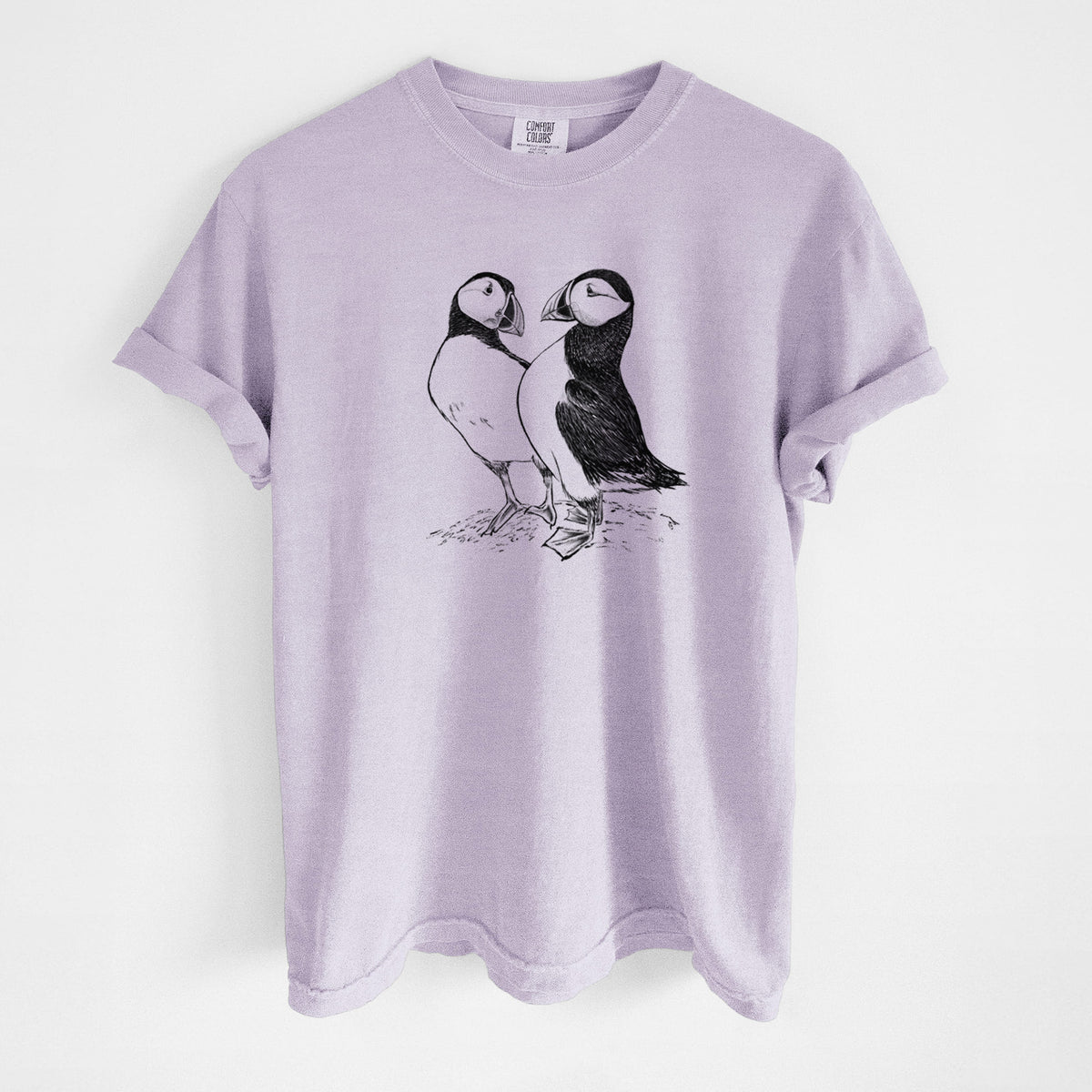 Orchid purple Comfort Colors t-shirt laid flat, printed with a detailed black ink illustration of two Atlantic puffins standing side by side.