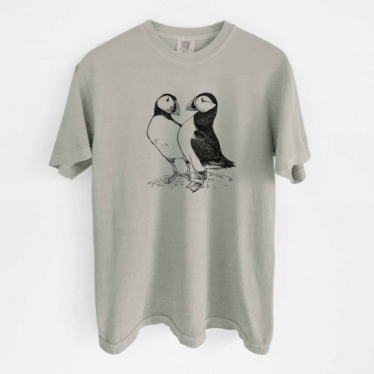Sandstone beige Comfort Colors t-shirt laid flat, printed with a detailed black ink illustration of two Atlantic puffins standing side by side.