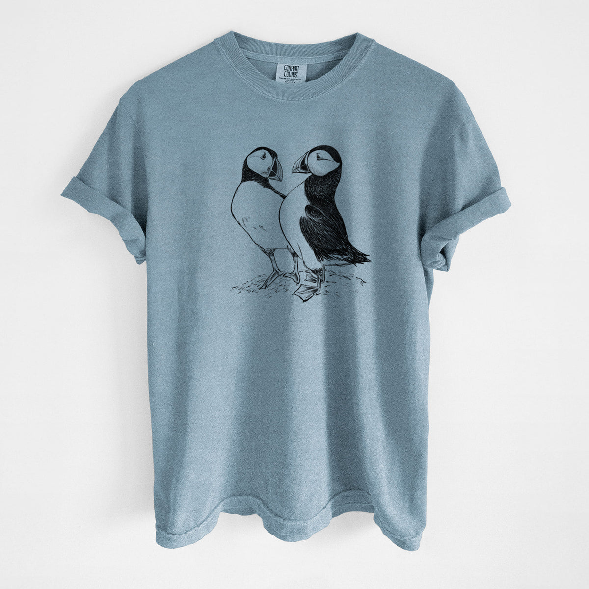 Steel blue Comfort Colors t-shirt laid flat, printed with a detailed black ink illustration of two Atlantic puffins standing side by side.