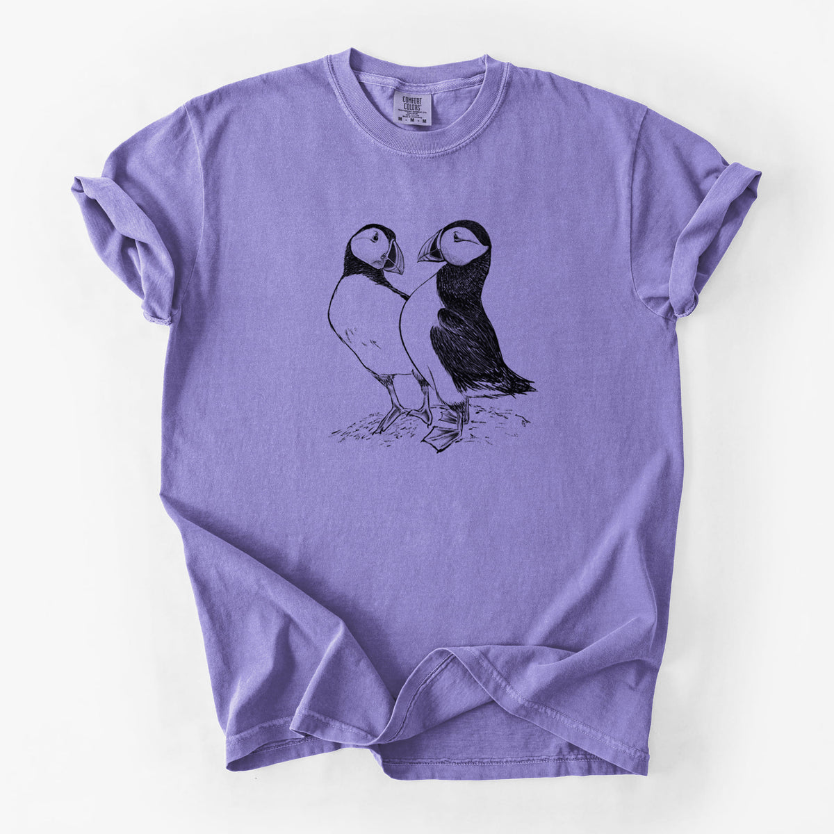 Violet purple Comfort Colors t-shirt laid flat, printed with a detailed black ink illustration of two Atlantic puffins standing side by side.