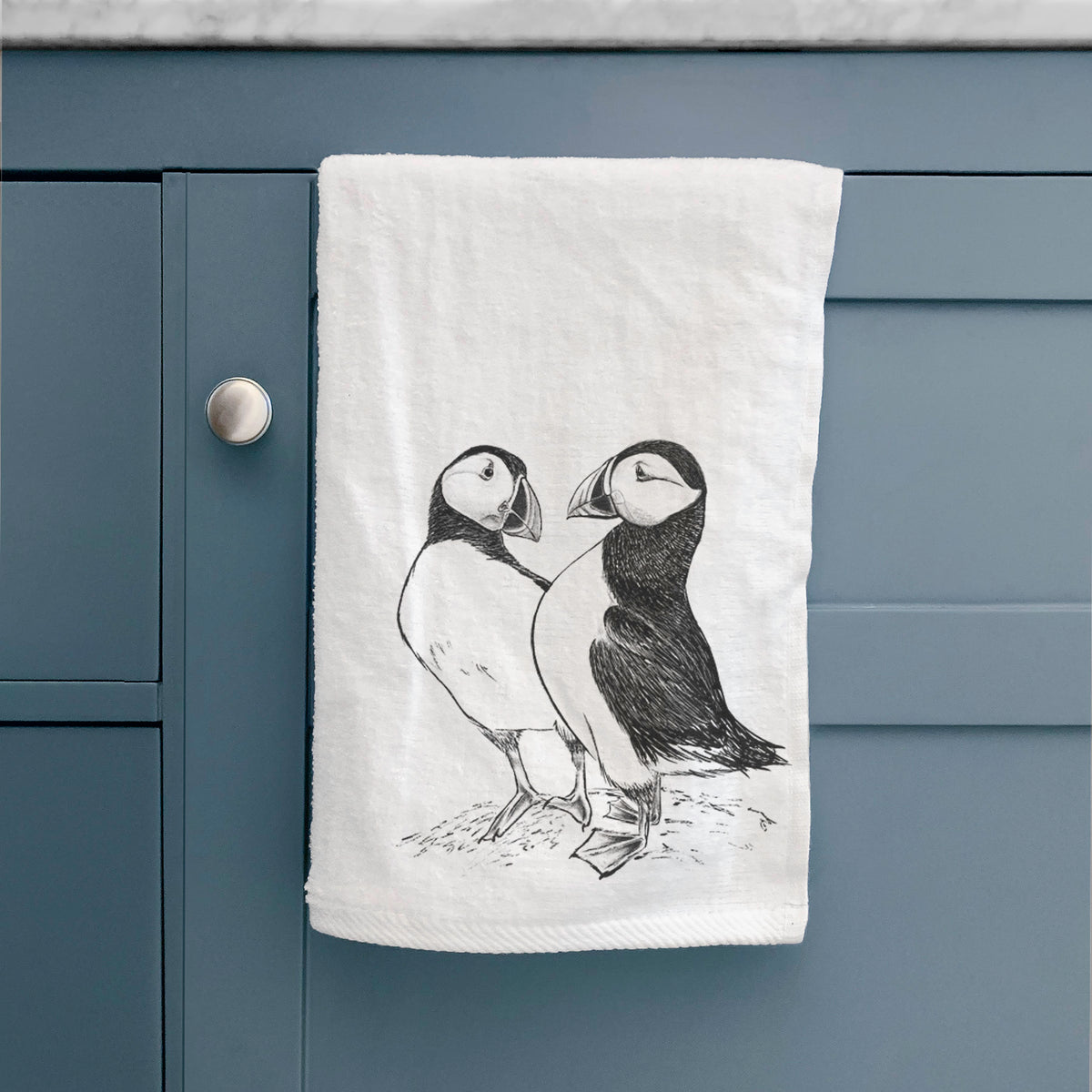 White hand towel printed with a detailed black ink illustration of two Atlantic puffins standing side by side, hanging on a cabinet door.